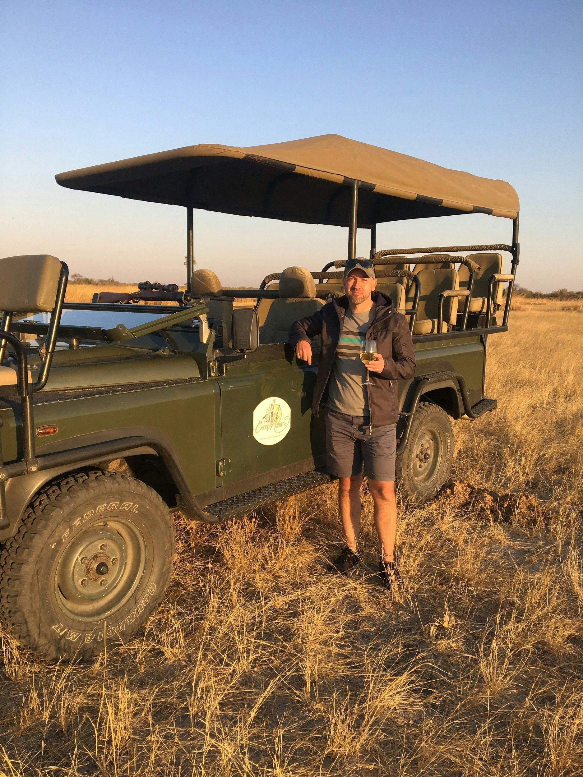 A white man holding a glass of wine stands beside a 4WD in open savannah
