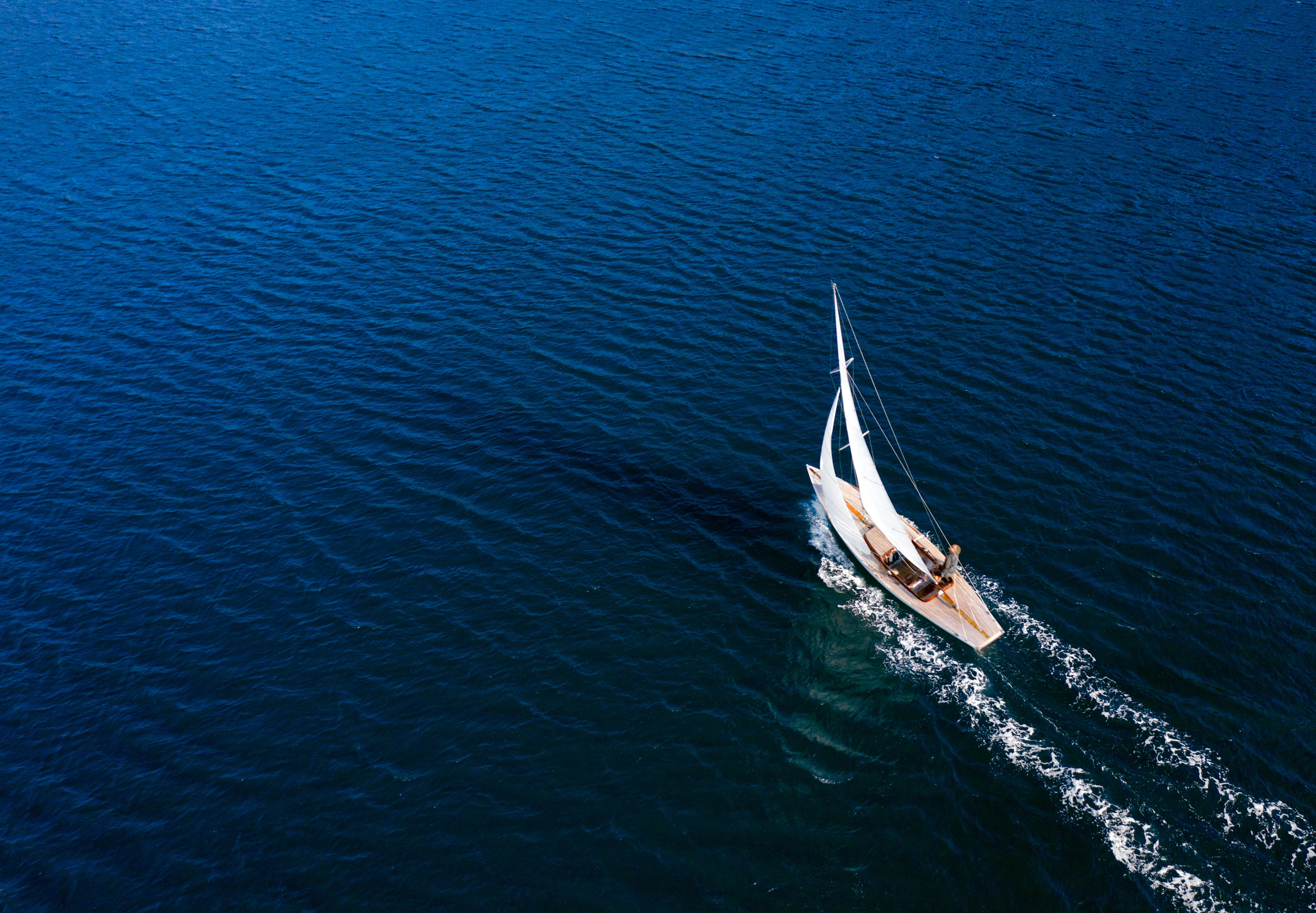 An aerial view of a small sail boat in a big patch of blue ocean. A man is visible sailing the boat.