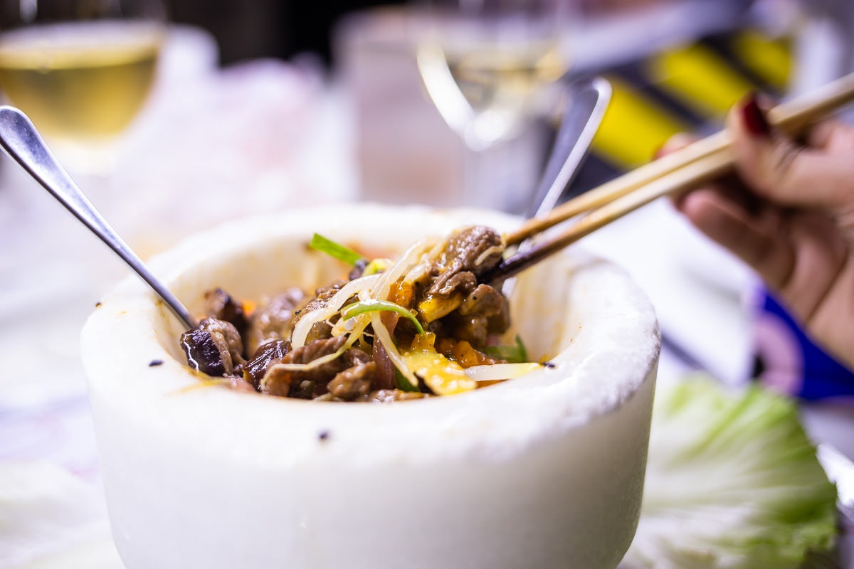Close up of a sturdy white bowl, containing beef and vegetables. A diner's hand is in soft-focus and they are using chopsticks to hold up some of the beef.