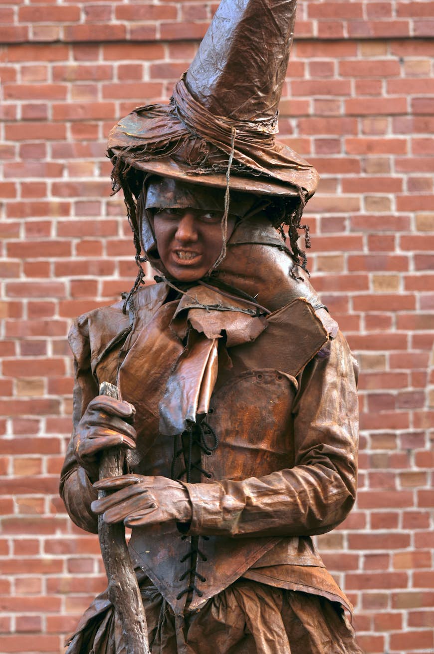 A woman dressed in dark brown leather witch garments holds a wooden staff in front of a brick wall 
