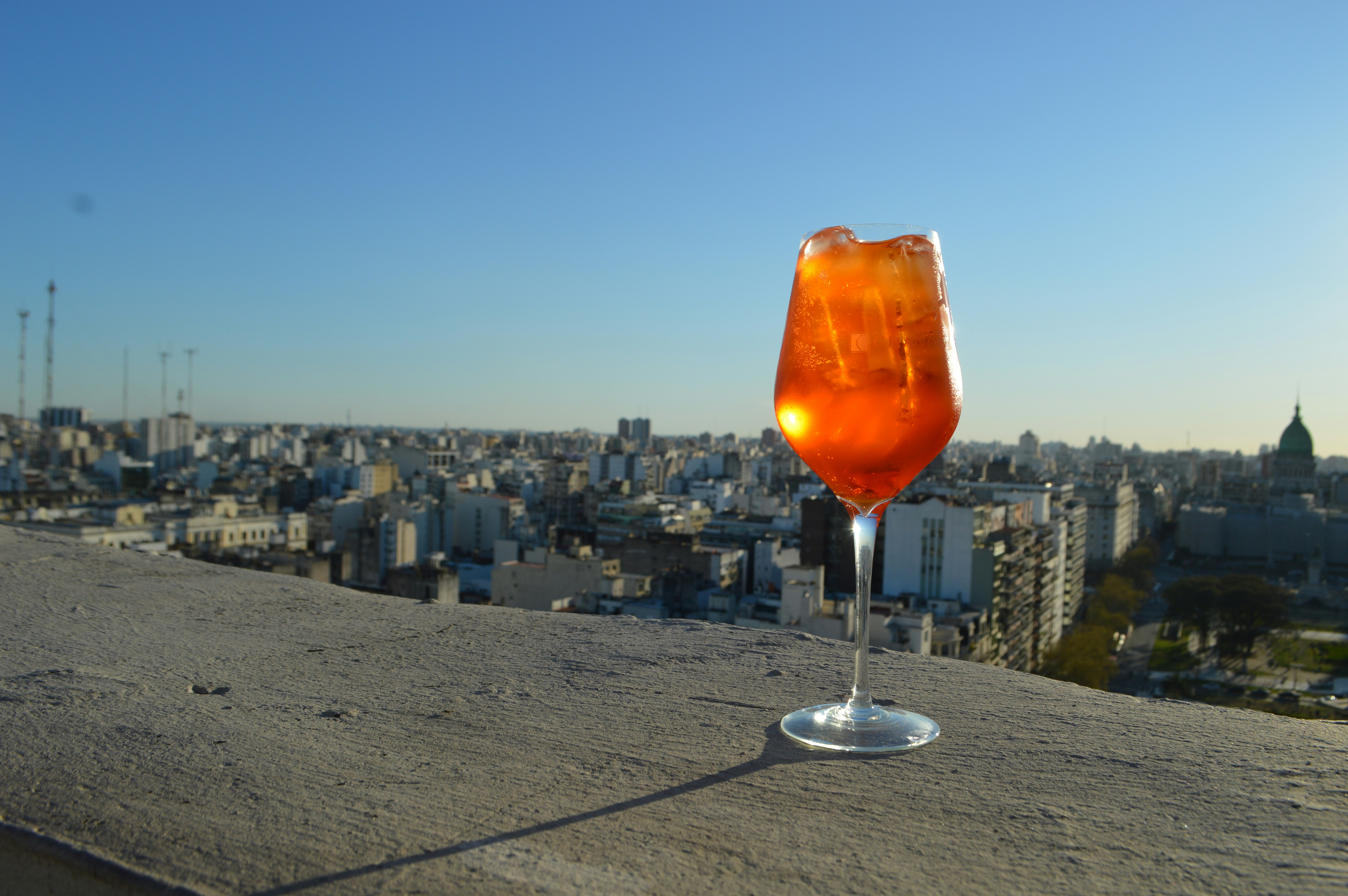 A wine glass filled with an amber-colored liquid rests on the stone ledge of a rooftop bar overlooking the Buenos Aires skyline