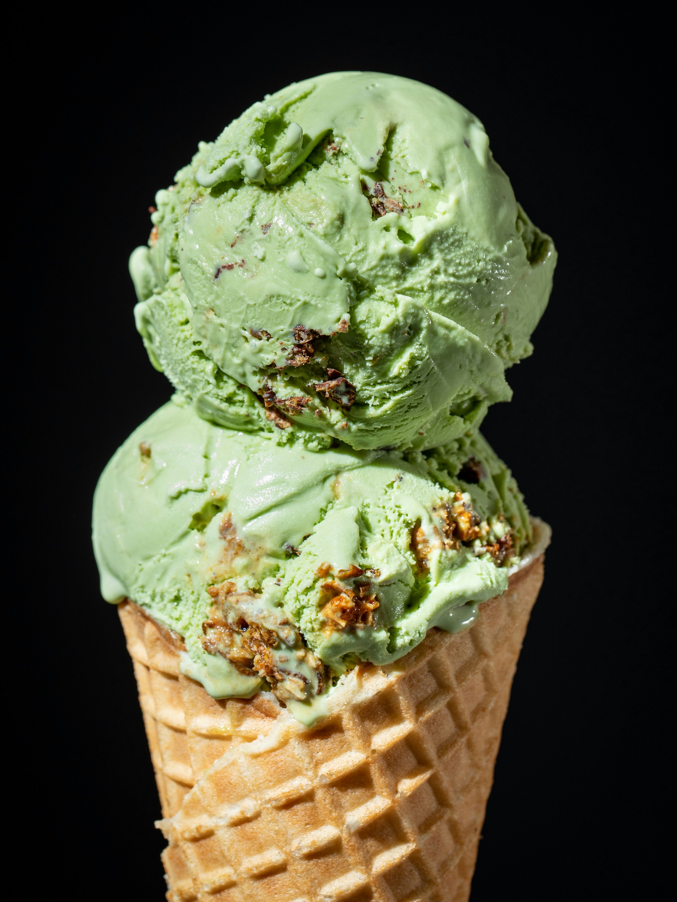 Two scoops of green-colored ice cream on a waffle cone. If you look closely, you can see chunks of chocolate-covered crickets. 