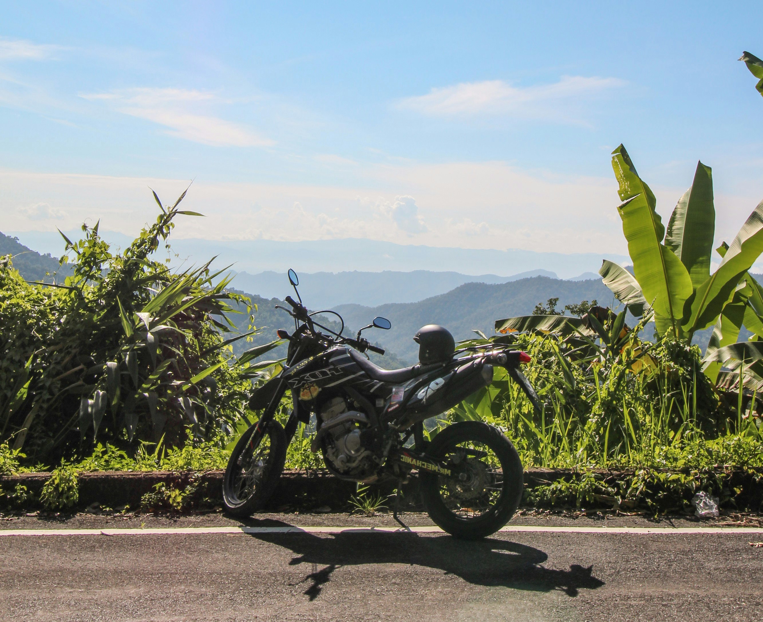 A motorbike is parked at a scenic viewpoint along the Samoeng Loop near Chiang Mai. A helmet sits on the bike seat while views of green valleys are visible beyond.