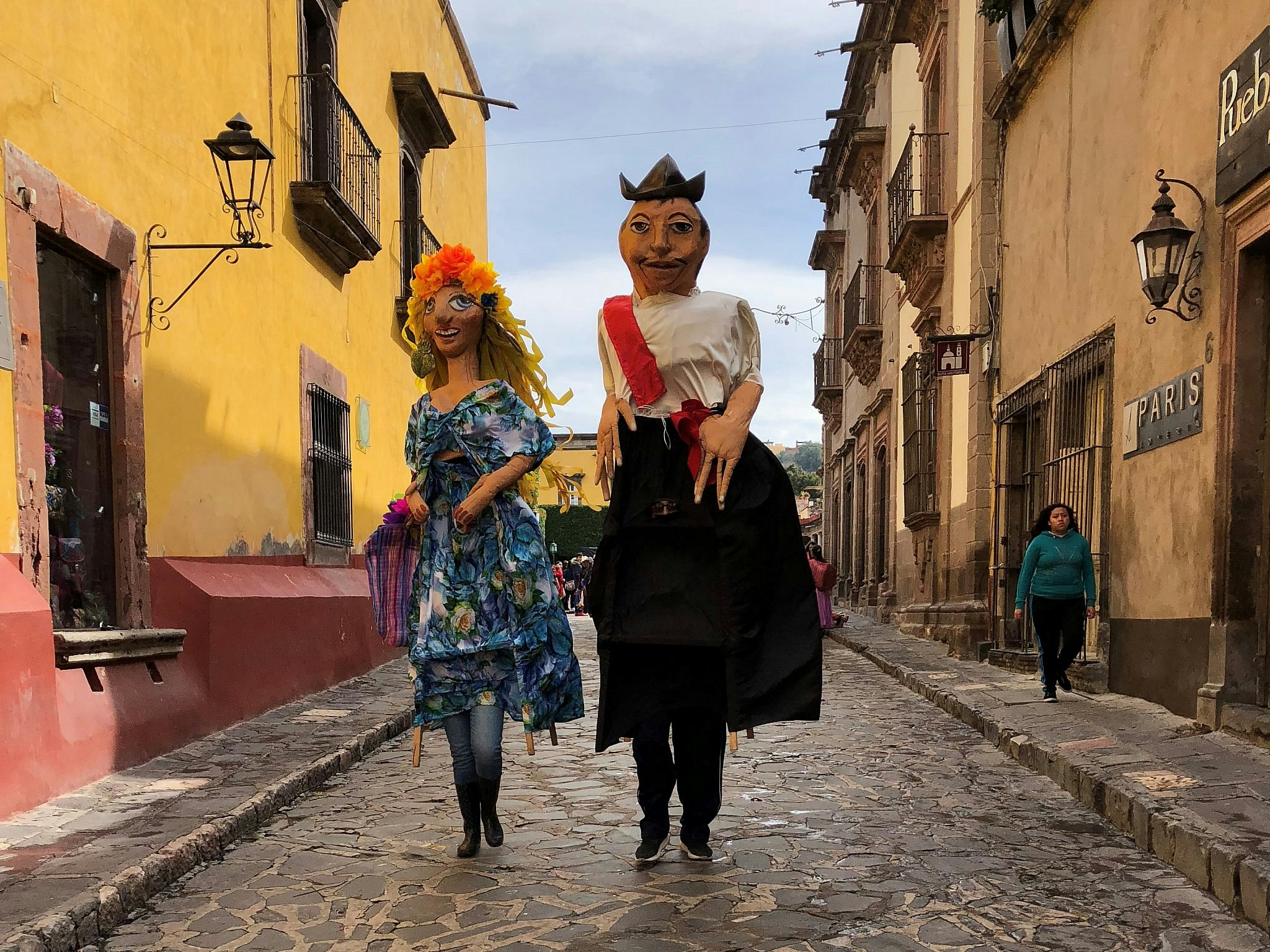 Two people dressed as giants with enormous paper mache heads walk down a street lined with colourful period buildings in San Miguel de Allende, Mexico.