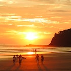 A group of surfers walk towards the water as sun creates a golden glow on the water and the sand