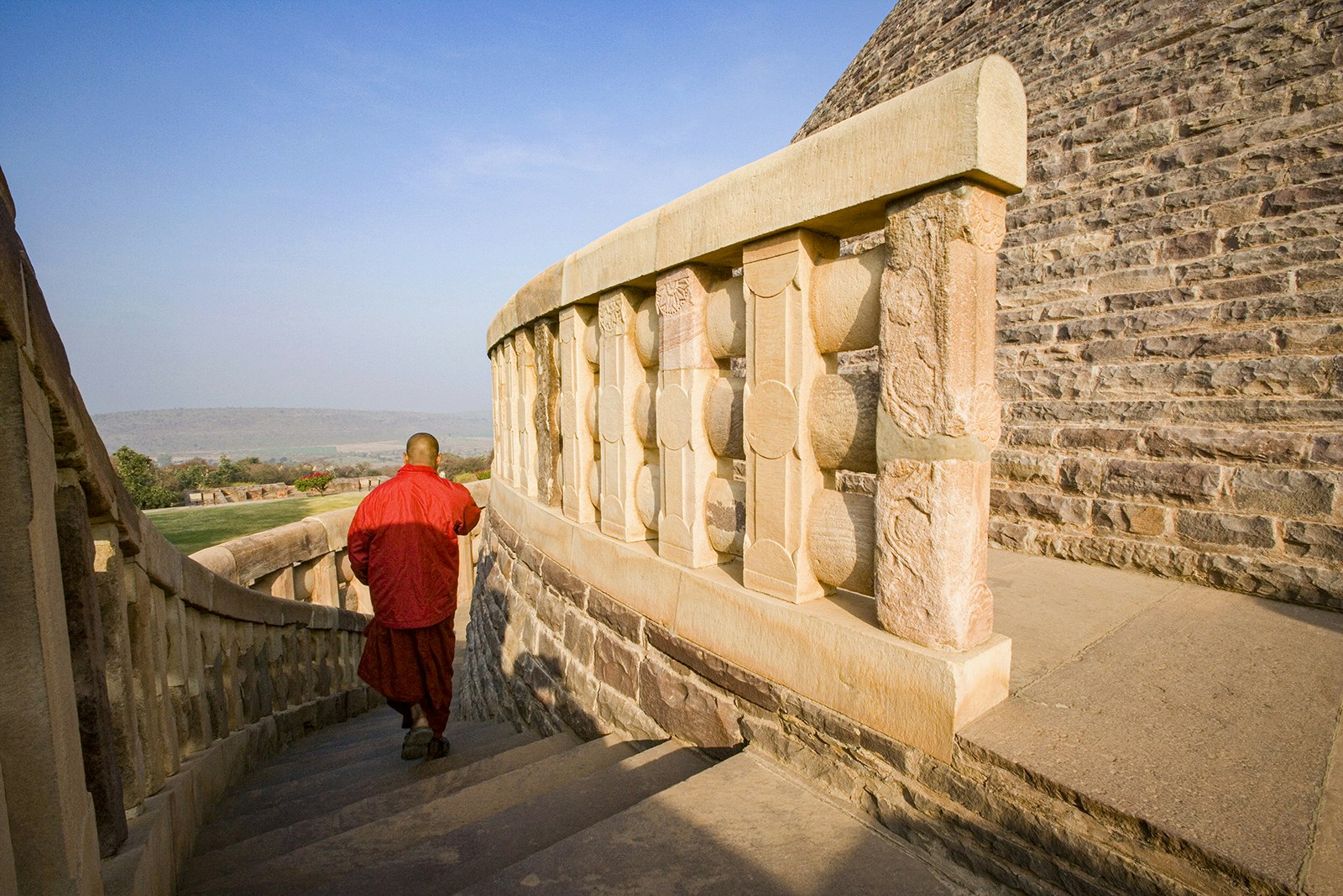 A monk clad in orange walks down a curved staircase encircling the rounded walls of the Great Stupa in Madhya Pradesh, India.