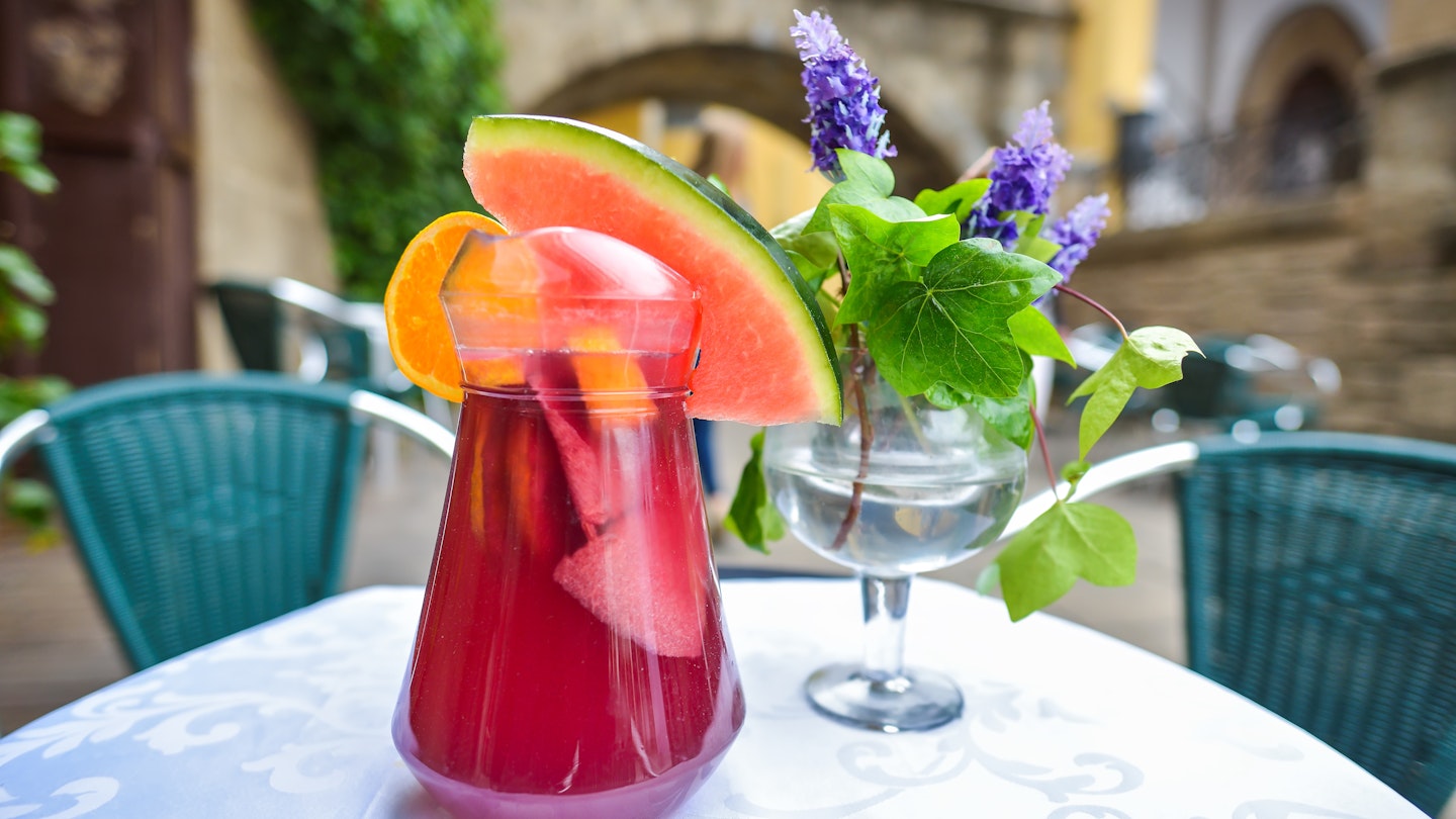 A large glass of ruby-colored Sangria. A large wedge of watermelon and orange slice hang off the rim of the glass.
