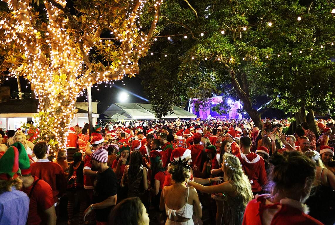 A huge crowd of people dressed as Santa in Wollongong, Australia, mingle under trees adorned with fairy lights at night time. There is a stage in the far distance and tented bars around the edges of the crowd.