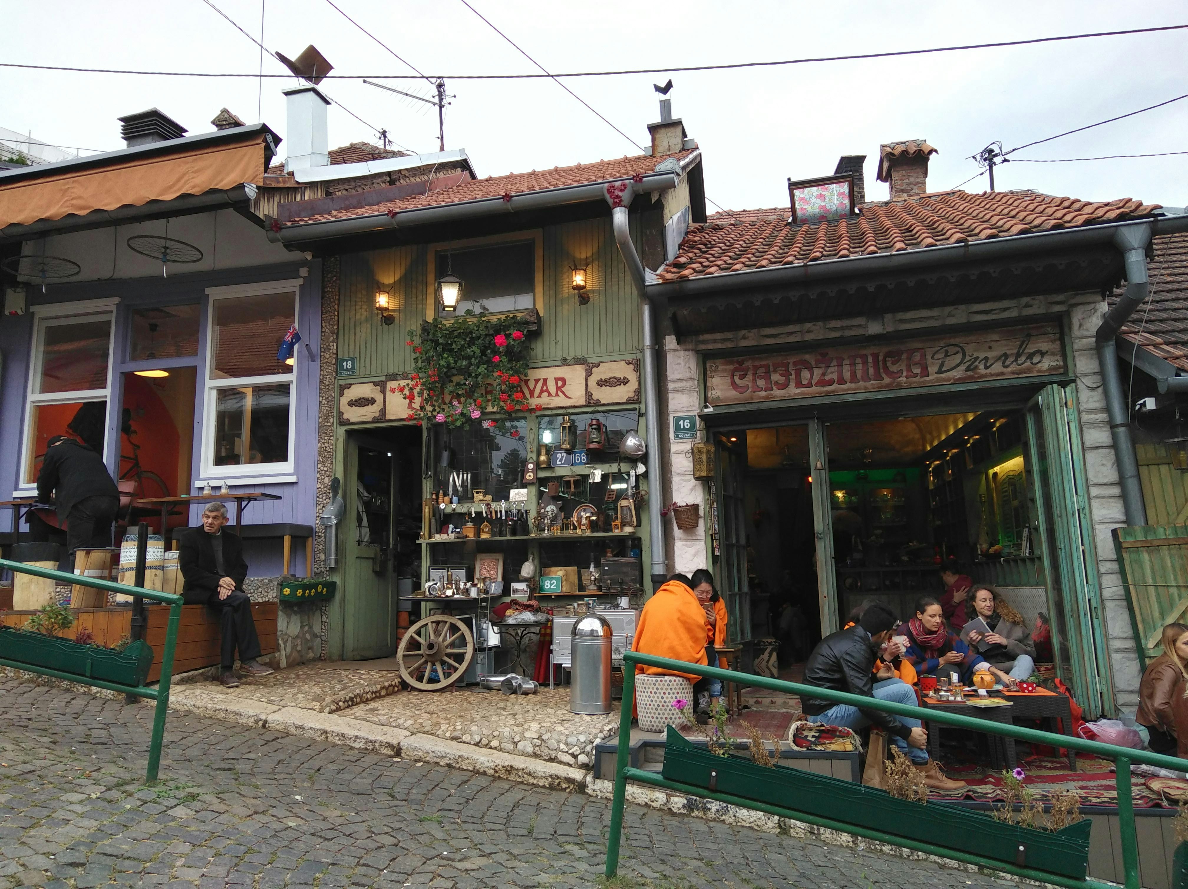 A group of people are sitting on tables outside a cafe on a quaint, hilly, cobbled street in Sarajevo. 