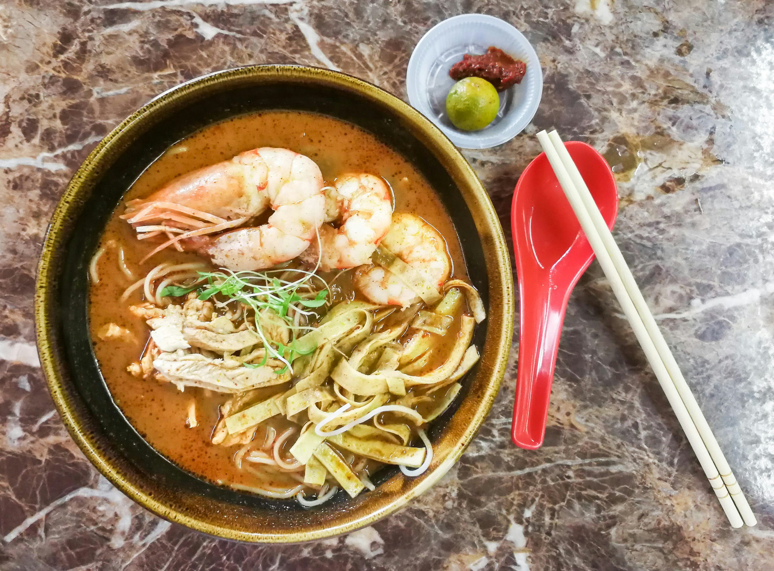 A bowl of Sarawak laksa as seen from directly above. The bowl is filled with a thick broth containing large noodles, strips of chickens and prawns.