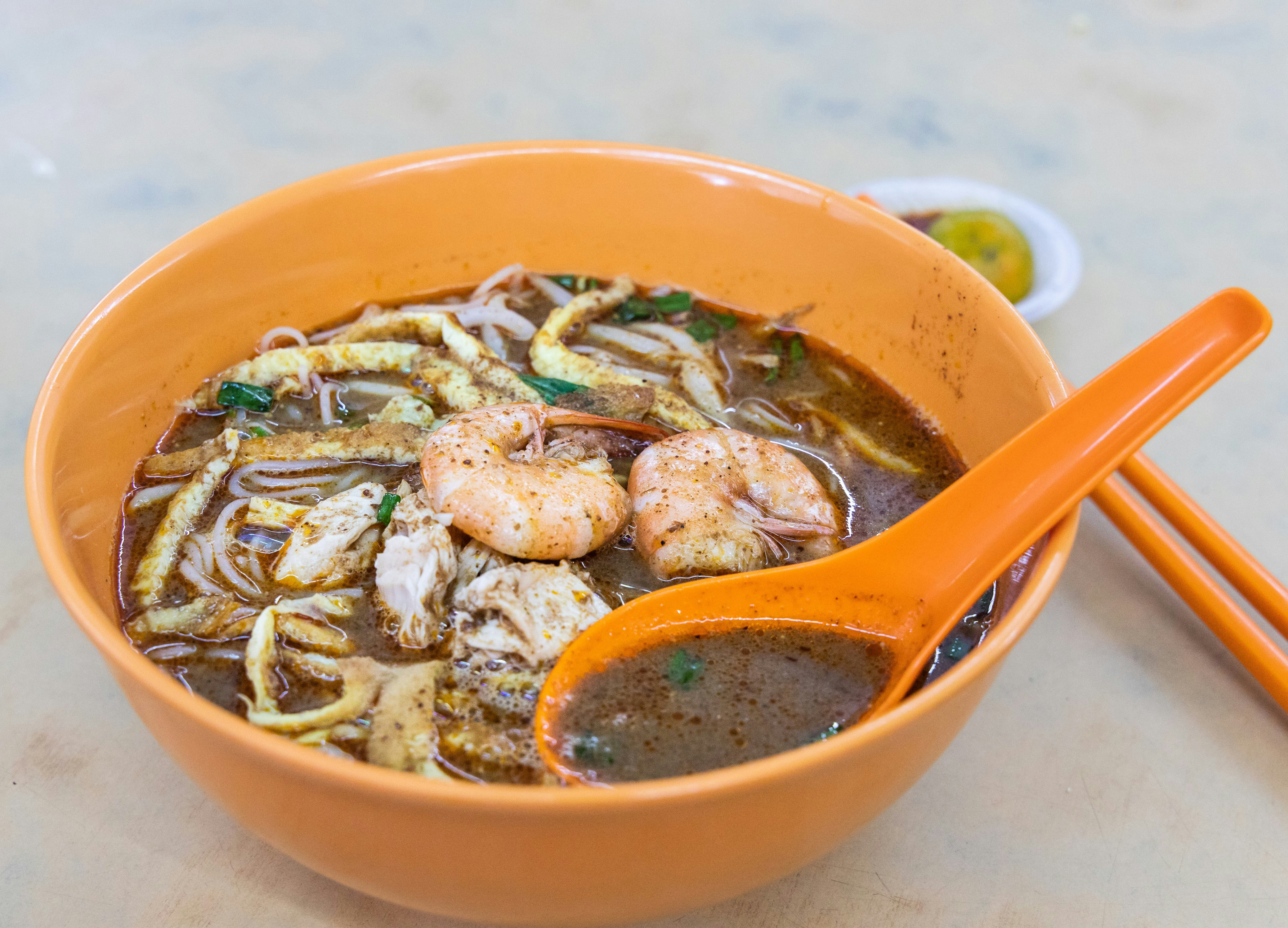 A close-up shot of Sarawak laksa in an orange bowl. The bowl is filled with a dark broth in which noodles, prawns and pieces of chicken are floating.