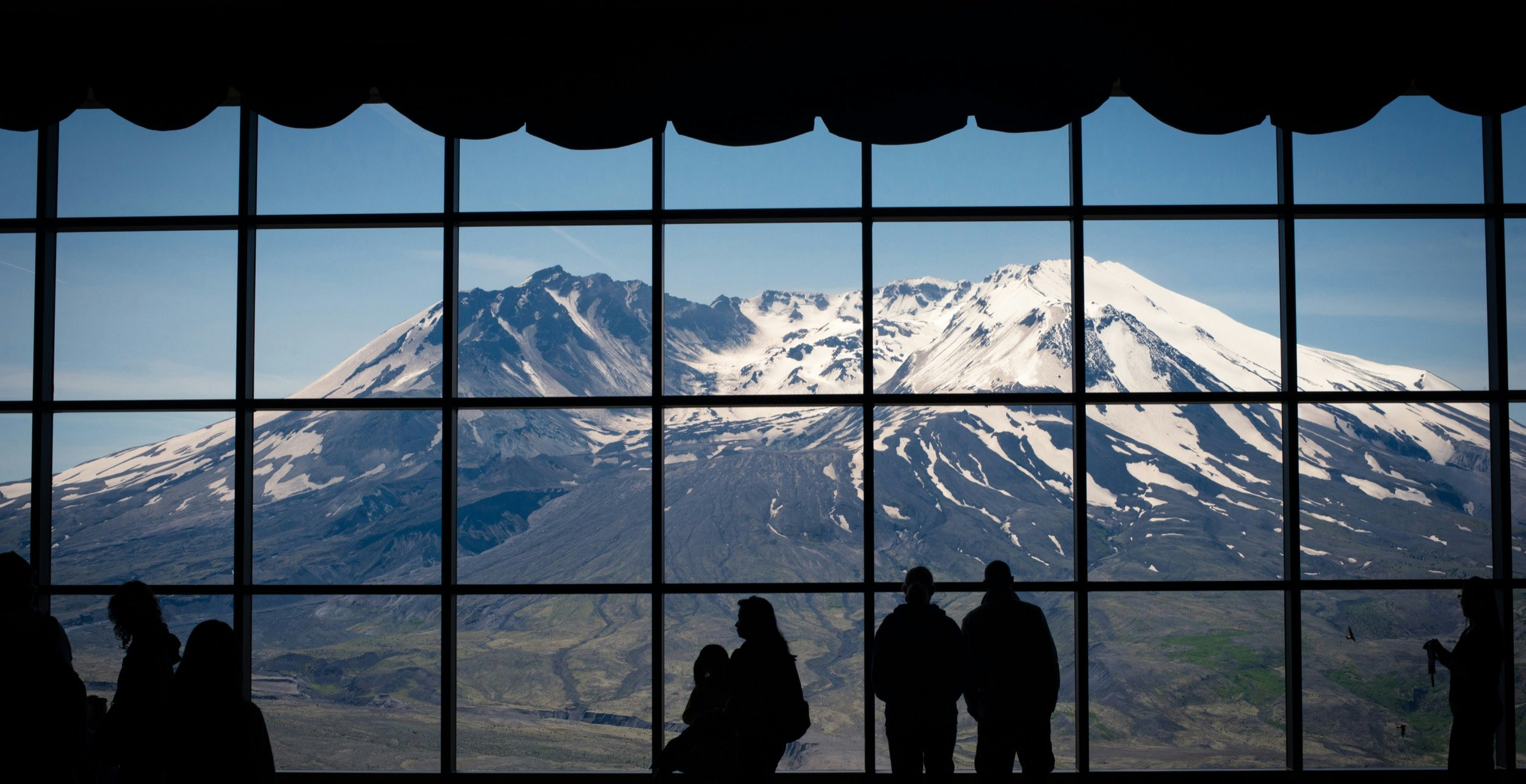 People stand in front of a large window looking out at the destroyed cone of Mt St Helens in Washington