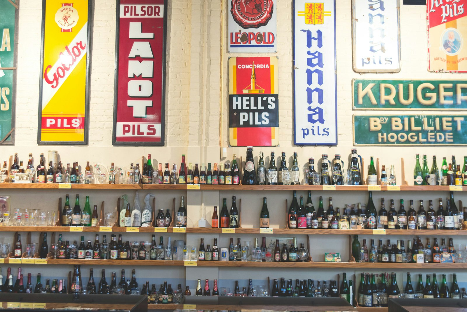 Wooden shelves are lined with beer bottles and glasses of different sizes and colours. Old-fashioned tin signs advertising beers are hung on the wall.