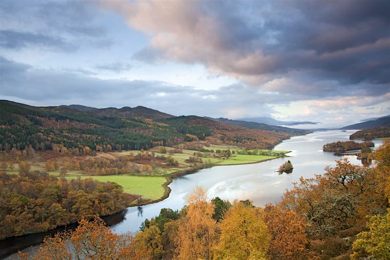 A Scottish loch is framed by rolling farmland and hills covered in a mixture of green and autumn-colored leaves.