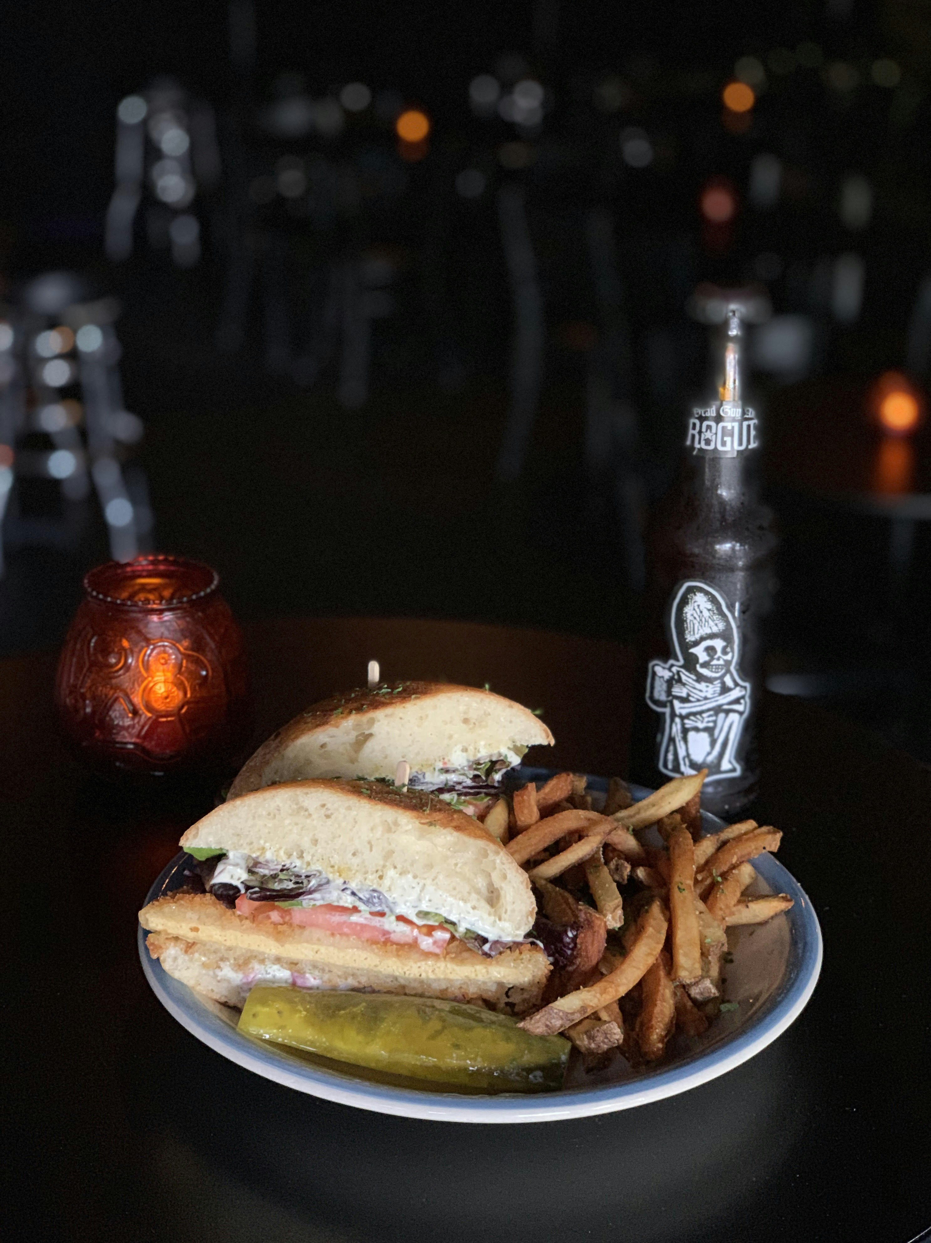 A plate filled with fries, a vegan sandwich and a pickle sits on wooden bar next to a beer bottle and a glass candle holder; Seattle vegan restaurants