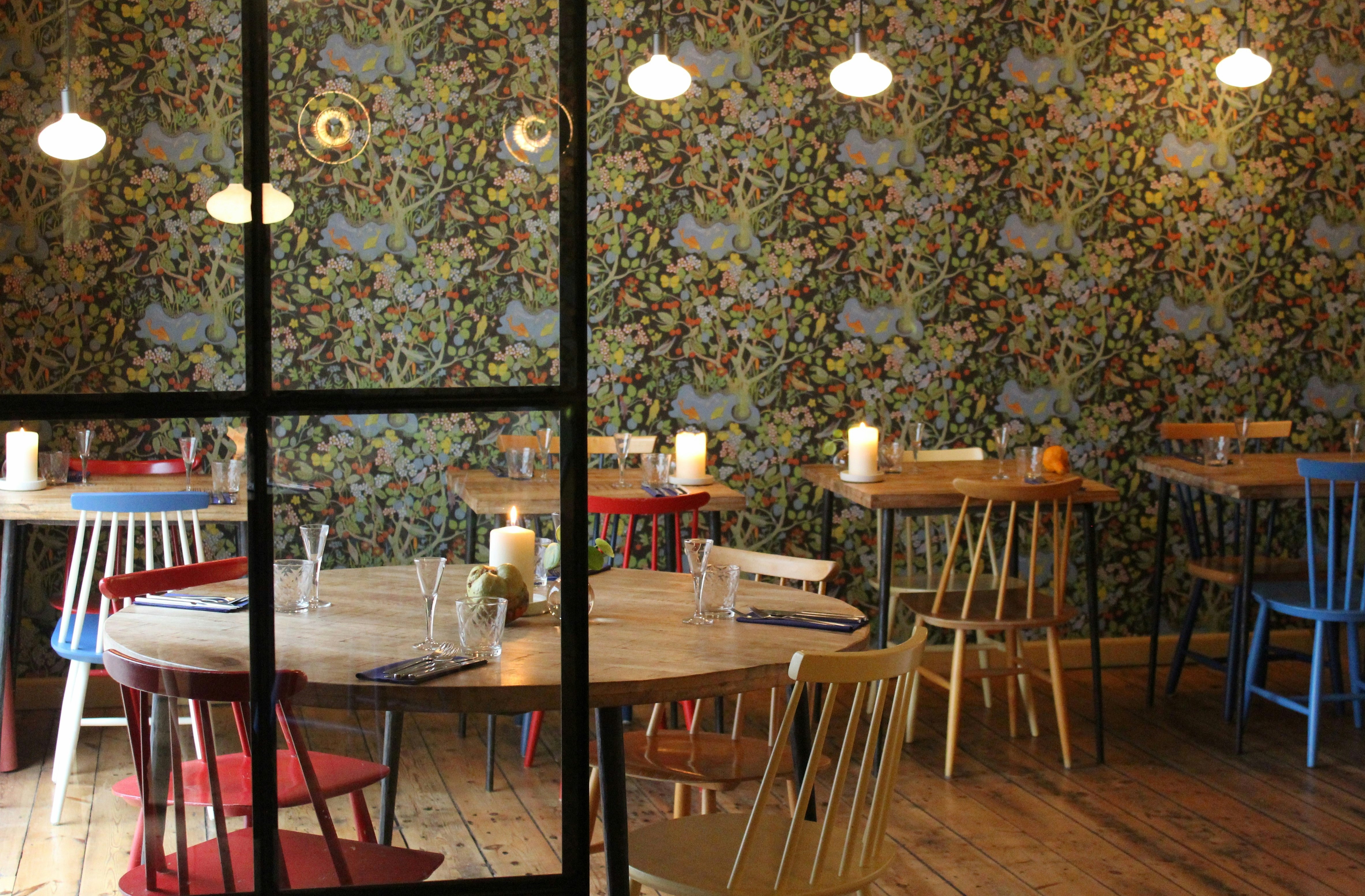 The interior of Selma restaurant in Copenhagen; it has floral wallpaper and wooden tables with mismatched chairs.