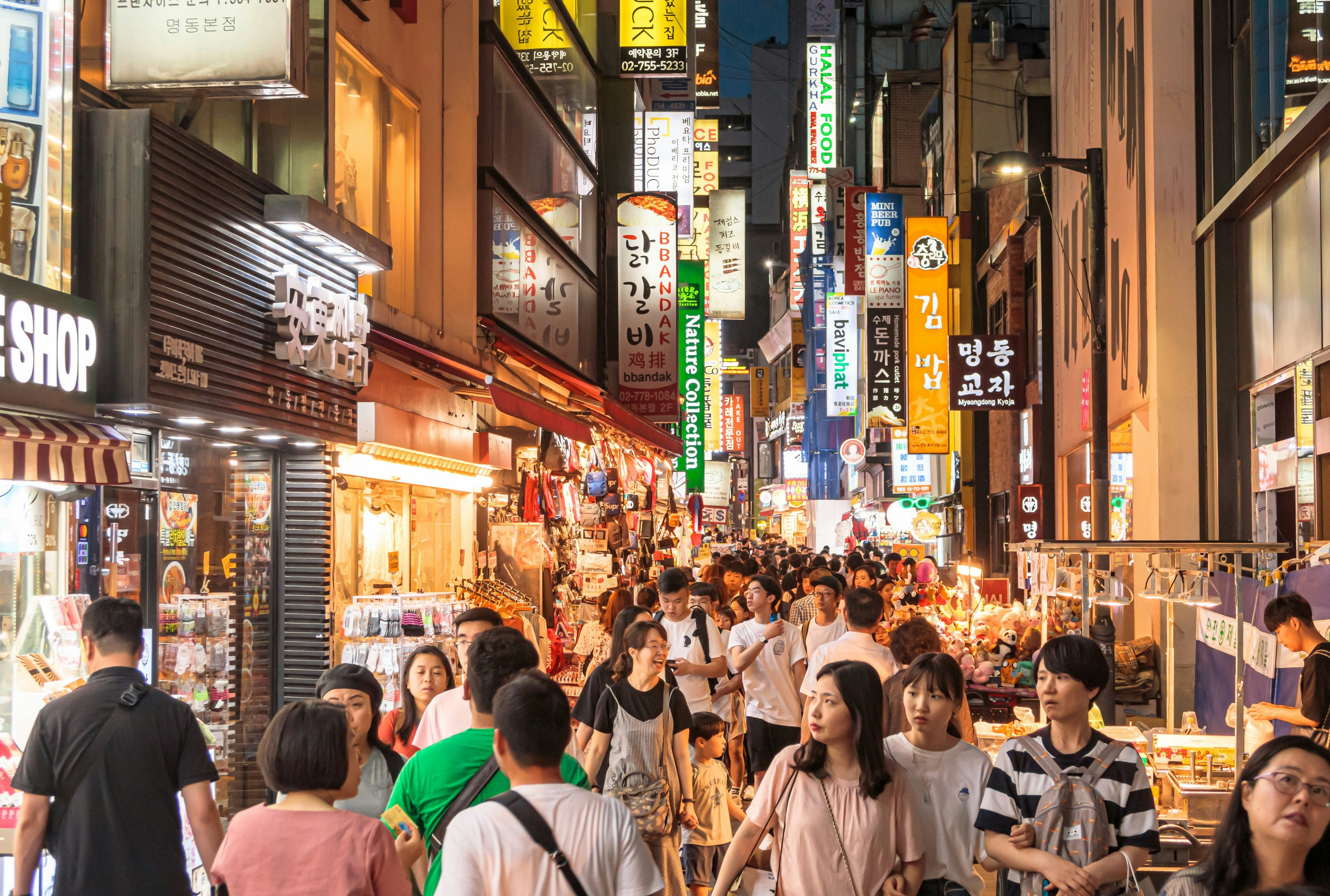 A crowded street in the Myeongdong district in Seoul. Walkers pass shops and stalls that flank either edge of the long street.