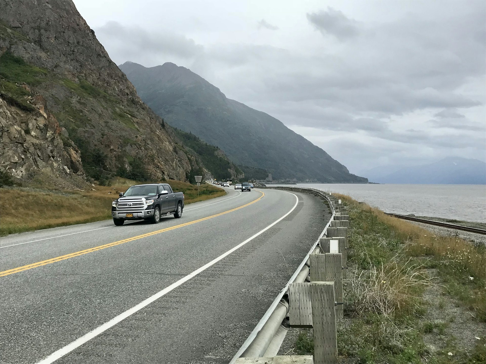 A pick-up truck drives towards the camera on a coastal highway, with the water to the sea on one side and rugged mountains on the other. 