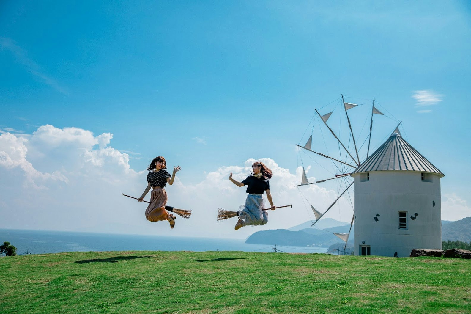 Two women pose for photos in Shodo-shima Olive Park by jumping up on brooms to look as if they're flying like the teenage witch in Studio Ghibli's film "Kiki's Delivery Service." A windmill sits in the background
