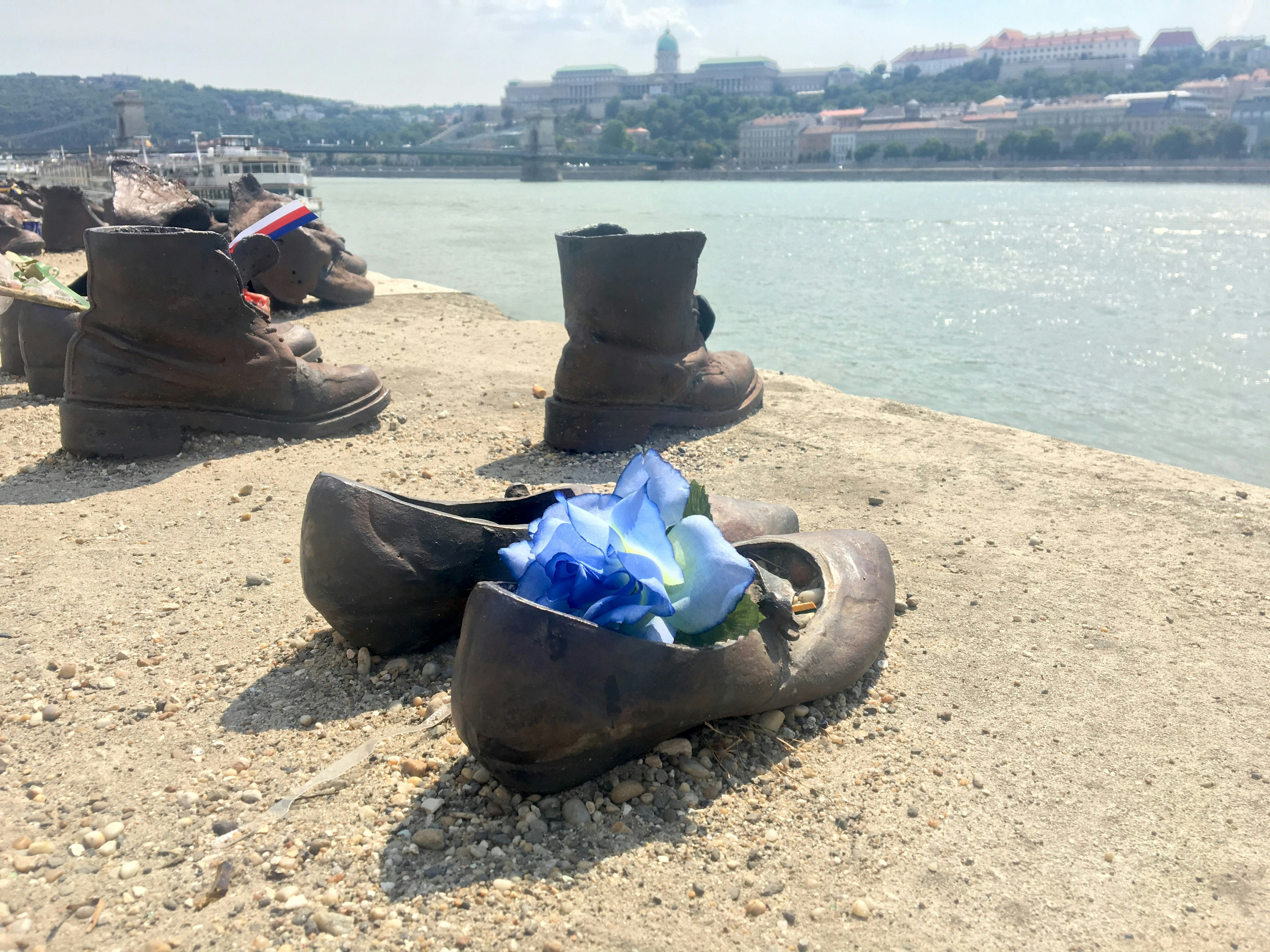 A metalic pair of women's shoes, which form part of the Shoes on the Danube Memorial, stand on the banks of the Danube River. The shoes have a flower placed in them. Across the river Buda Castle is visible.