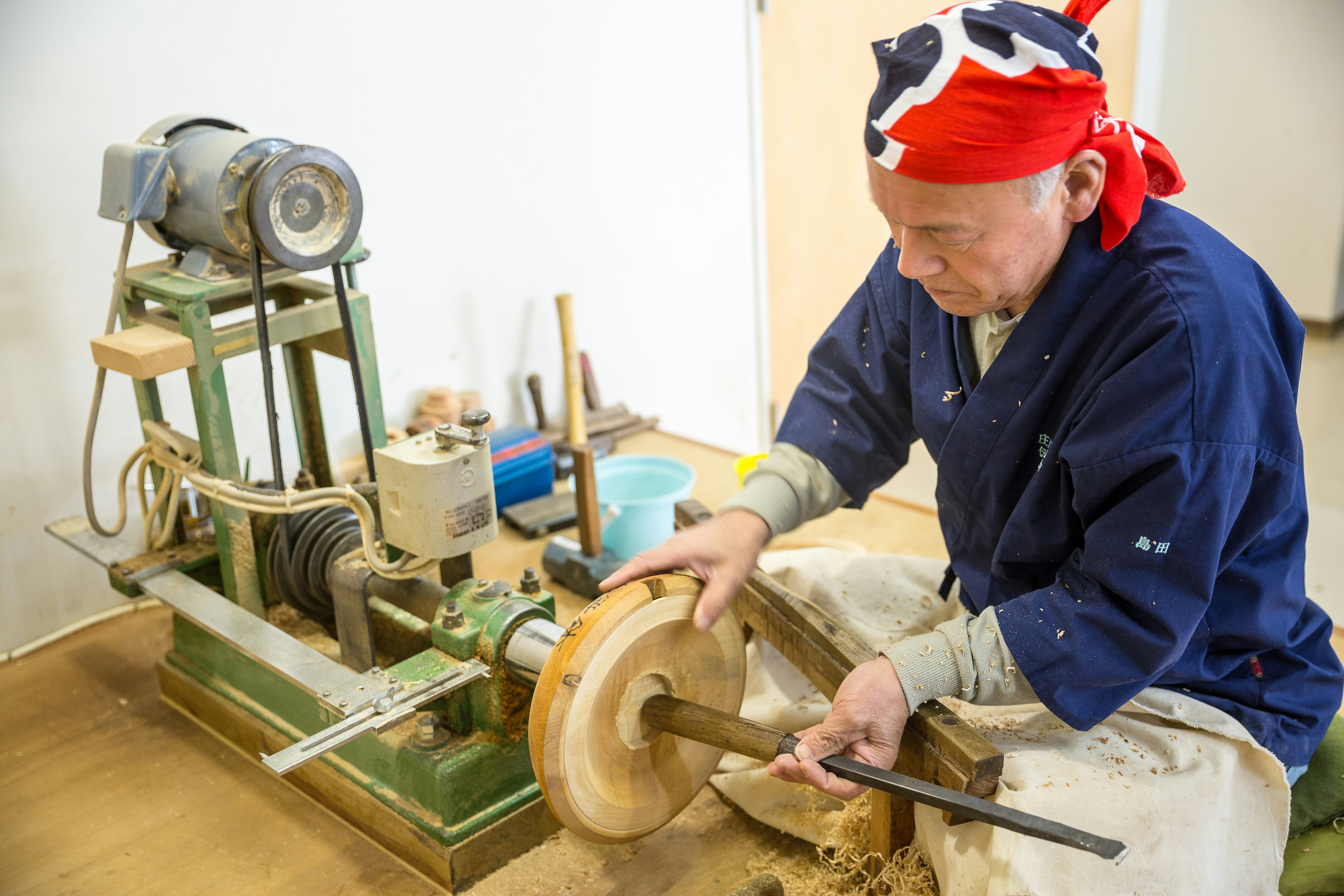 A craftsman wearing blue overalls and a bandana leans over some tools where the base of a bowl can be seen