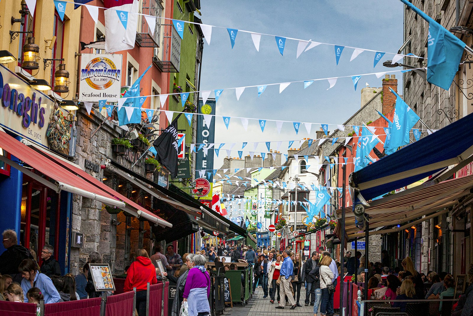 People walk down a crowded historic street lined with shops and bars. Pennants zig zag between the buildings above the street. Galway, Ireland.