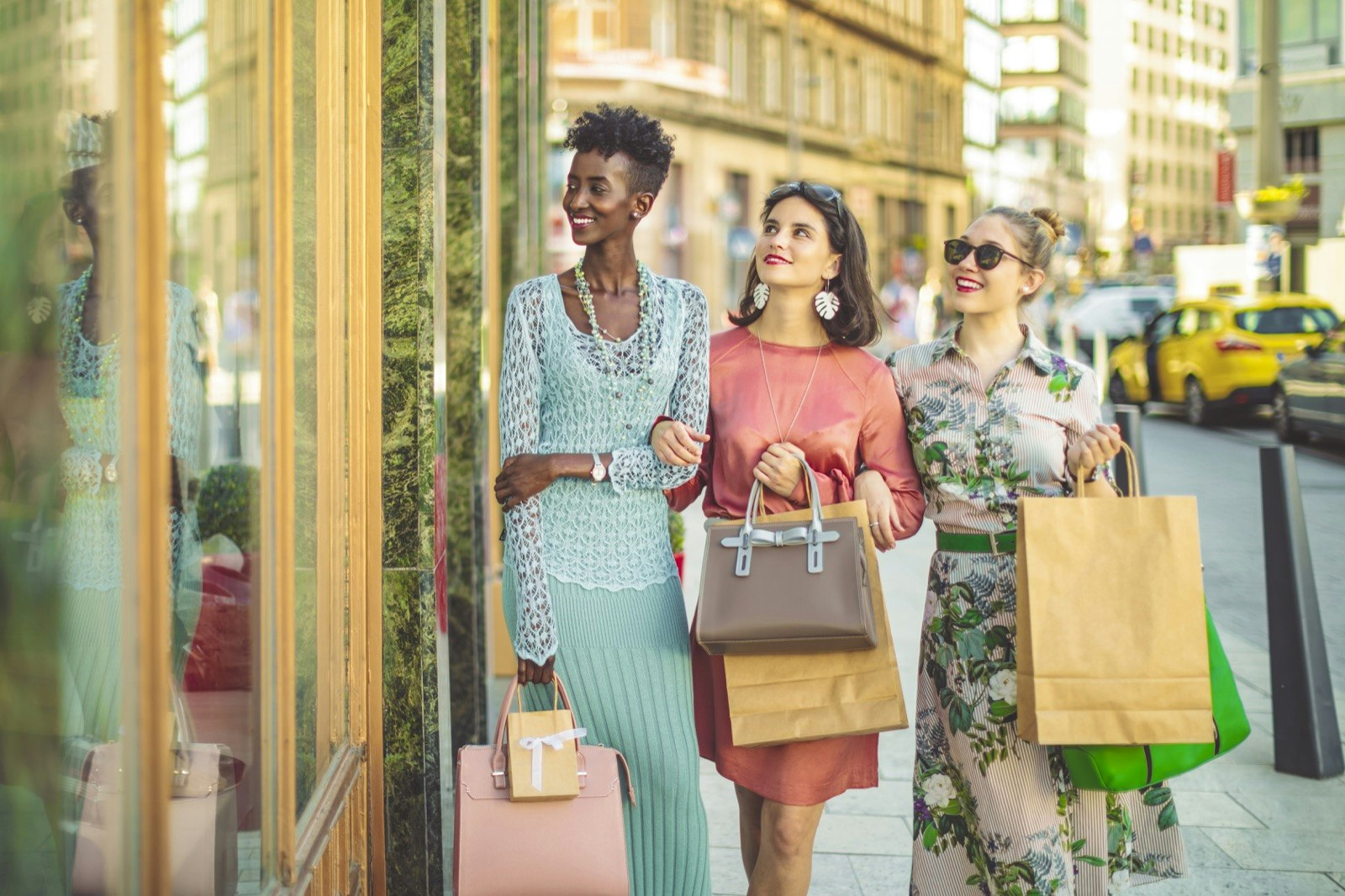 Three well-dressed women walk past a store window holding shopping bags and smiling