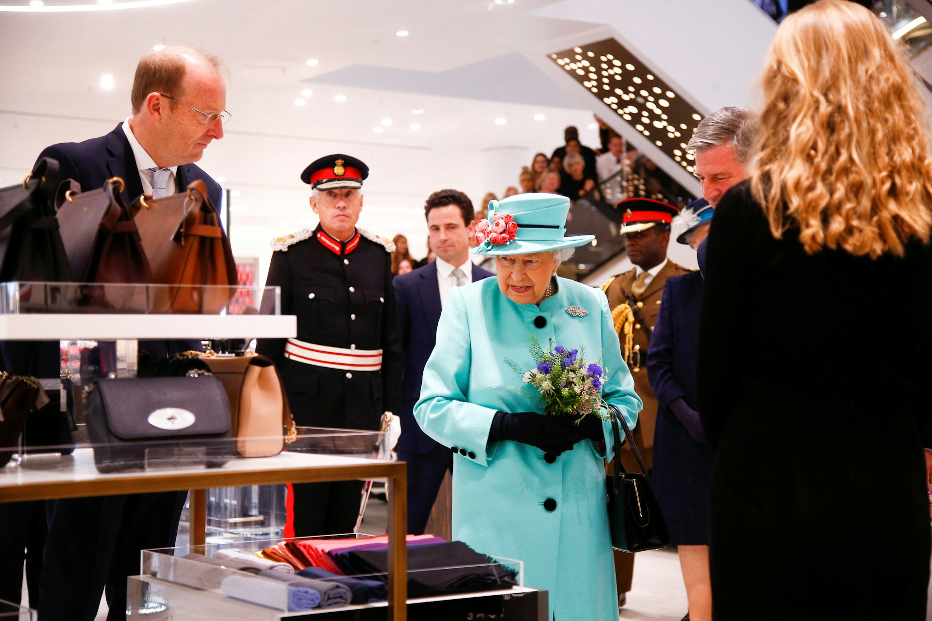 Queen Elizabeth II, wearing a blue coat and matching hat, looks on at handbags in a hightstreet retailer. 