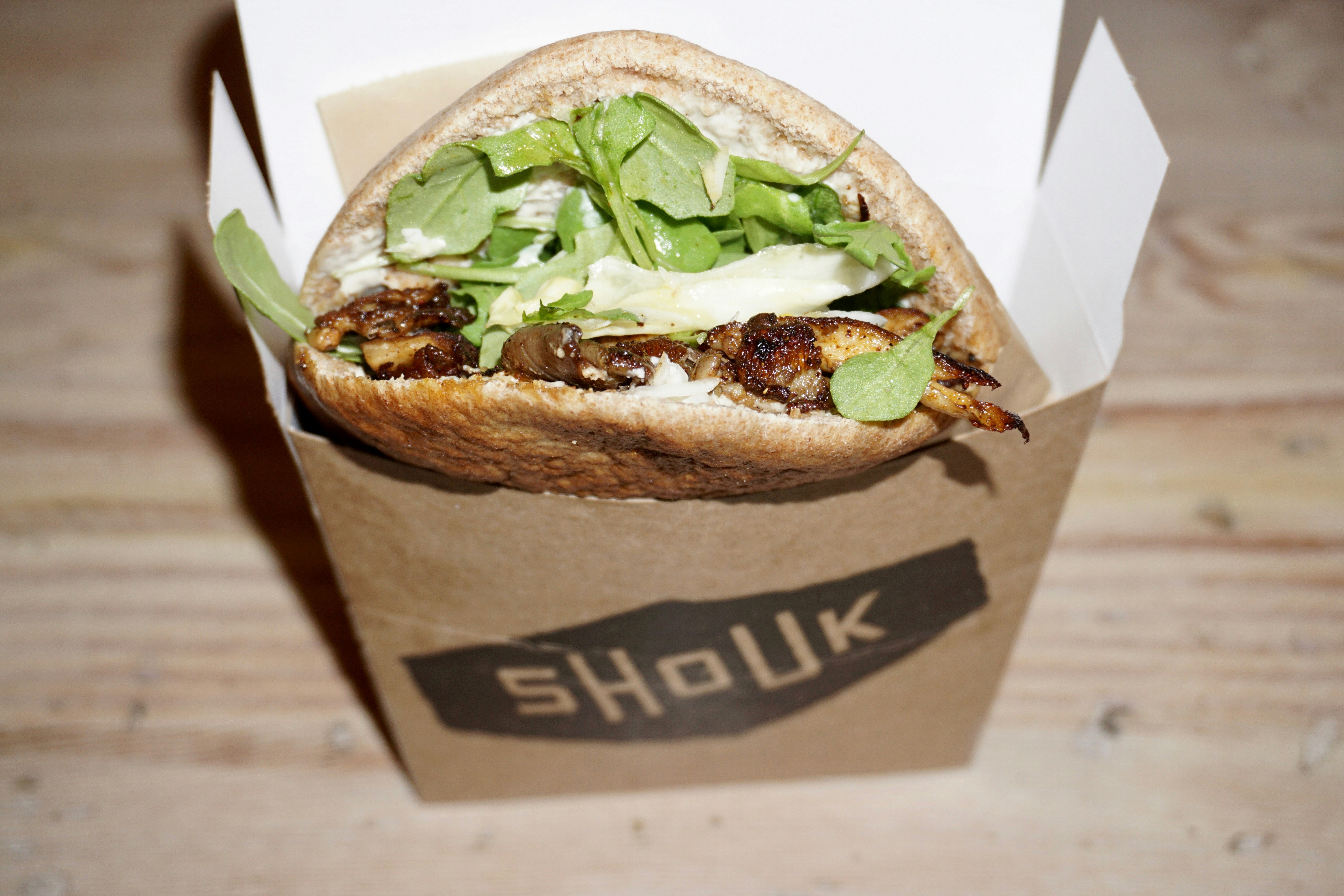 A cardboard box with "Shouk" printed on the front has a pita inside; it's stuffed with vegan ingredients such a mushrooms and green leaves.