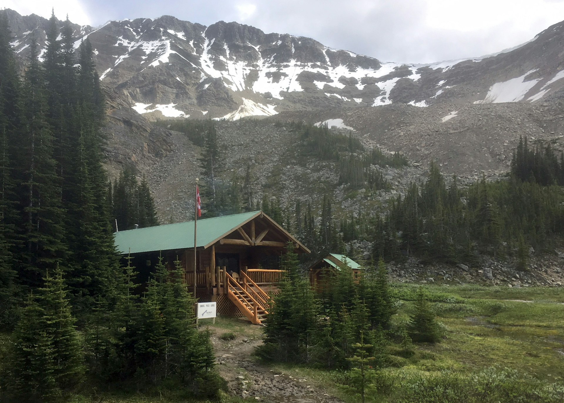 The Canadian flag flies next to a large lodge with a steep set of stairs and a snowy-in-spots mountain behind