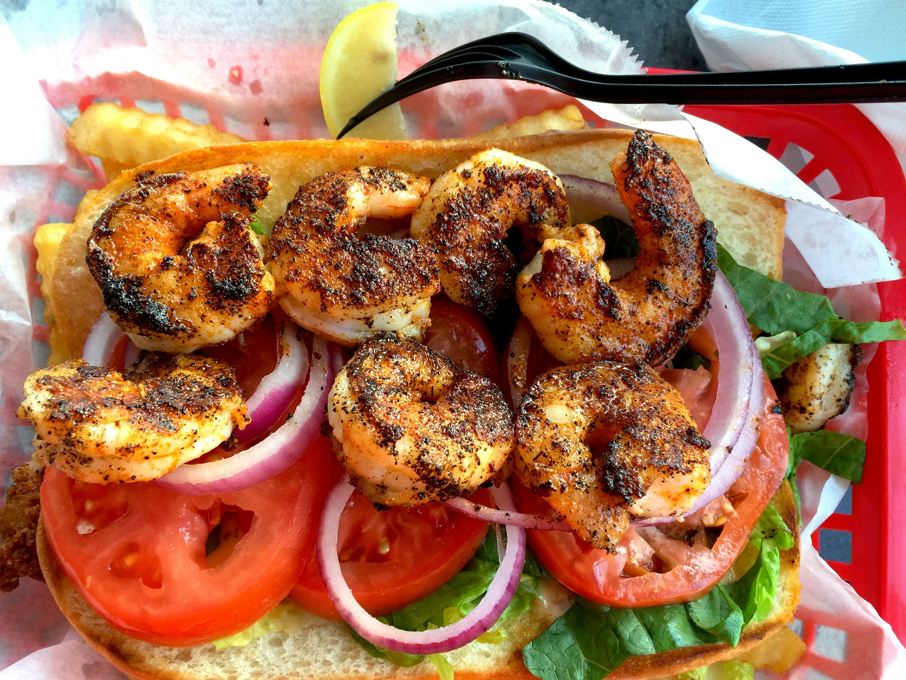 Grilled shrimp is placed on top of onions, tomato and lettuce to create an open-face sandwich. There are french fries under the sandwich and a black plastic fork resting on the red tray.