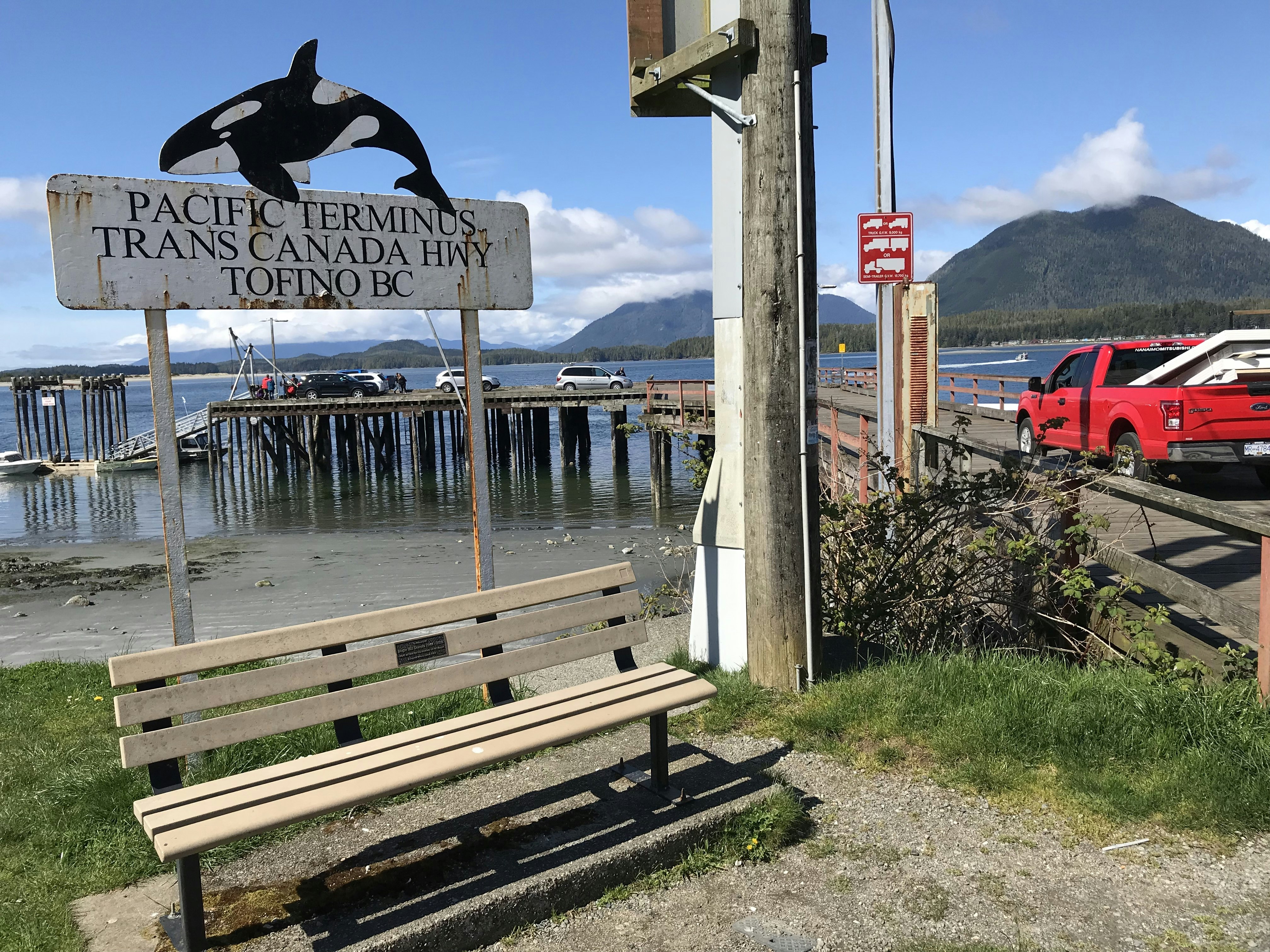A wooden bench sits with a white, worn metal sign behind it that reads "Pacific Terminus Trans Canada Highway Tofino BC" in a black serif font. An illustration of an orca is cut out of metal and hanging over the sign. In the background, several mini vans and SUVs in white and black pull onto a wooden pier from a steep ramp. A red pickup truck with white objects in the bed drives down the pier.