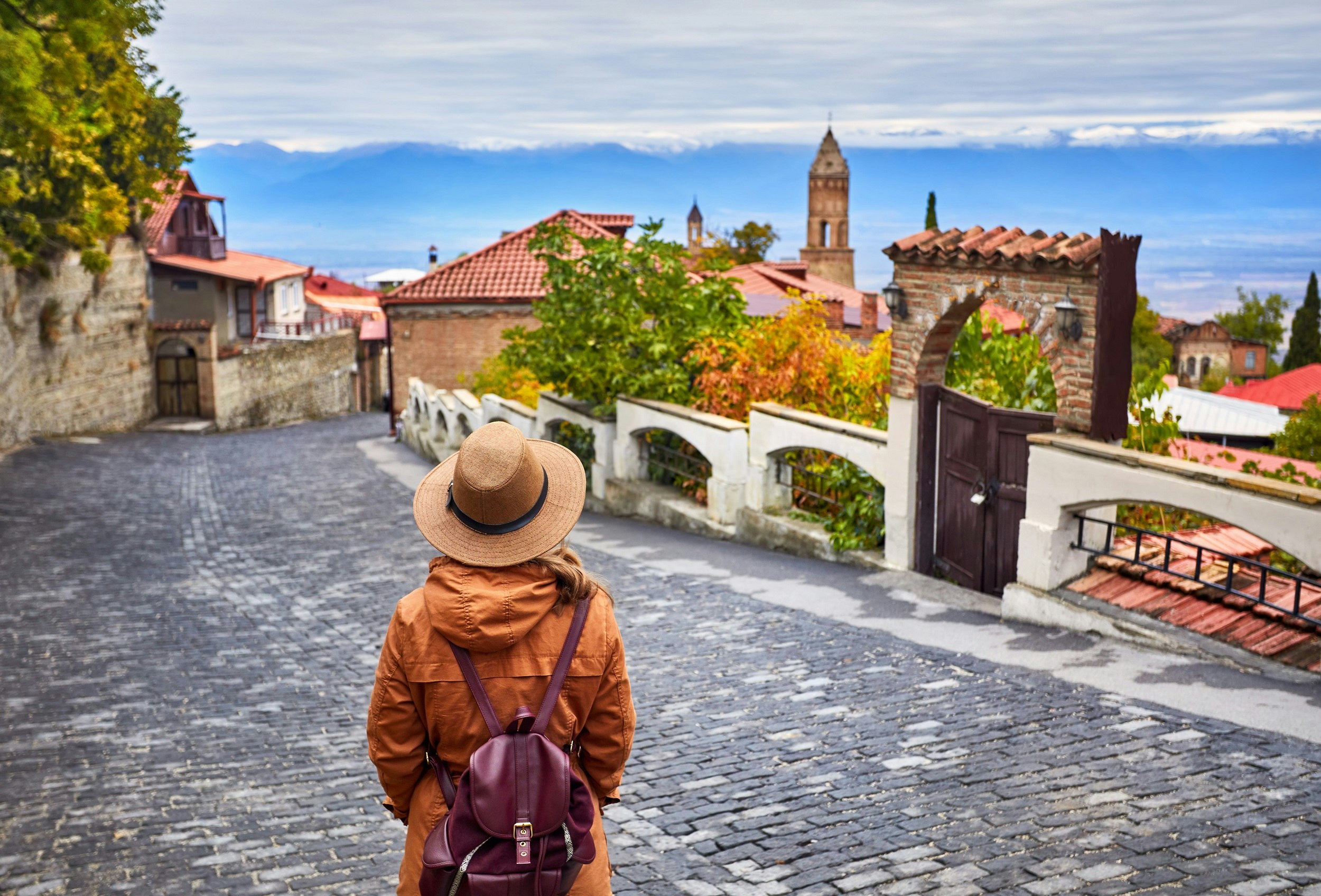 A woman walks down a wide cobbled street towards a town with a square church tower. Mountains are in the distance.
