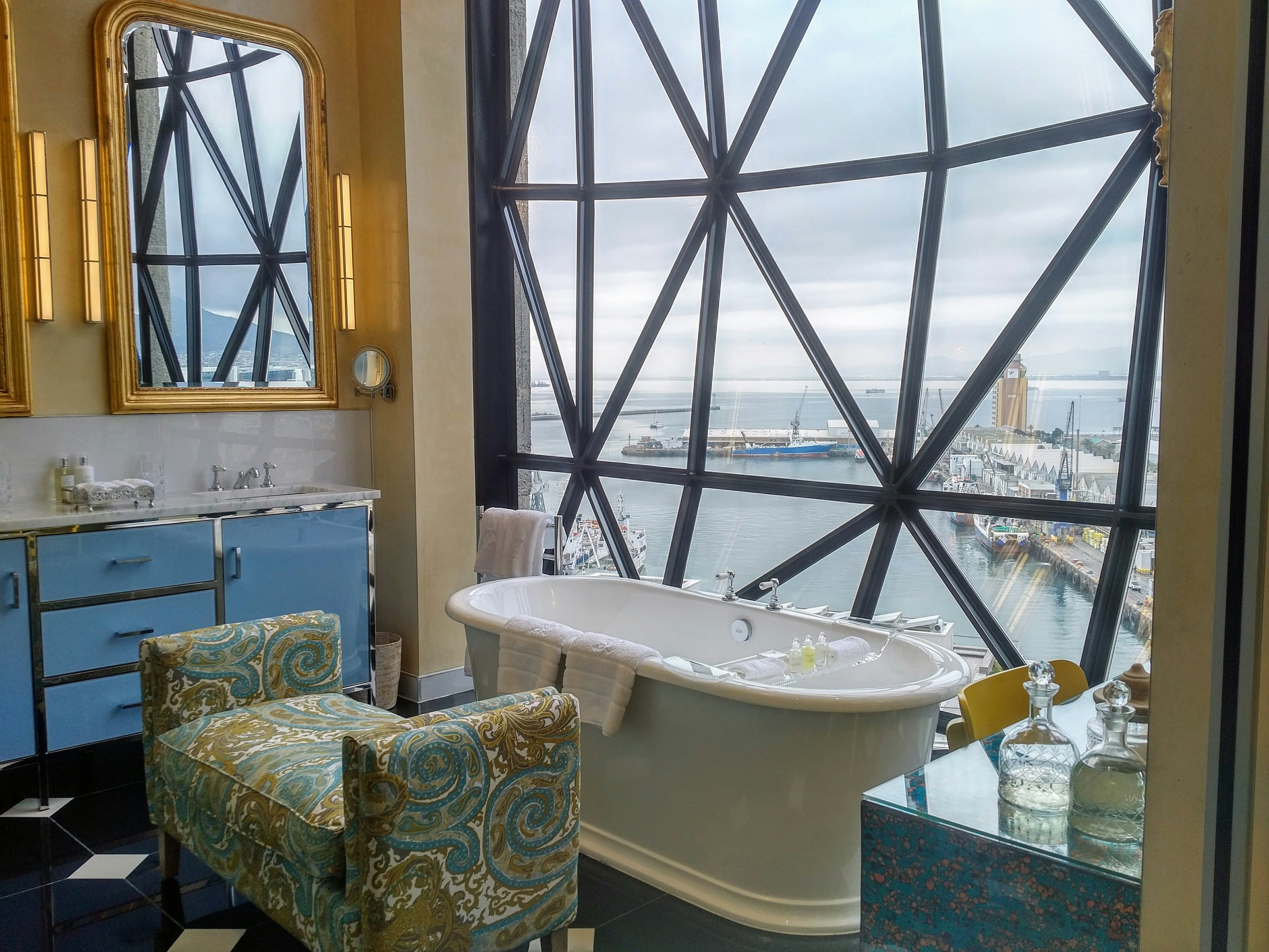 A white, vintage-style cast iron and porcelain soaking tub sits in front of an enormous window broken up into bold, geometric triangular panes. Parallell to the tub is a green and blue paisley Empire settee. Gold mirrors to the left reflect the window over a blue cabinet with a sink. In the foreground to the right are two crystal decanters filled with a pale liquid.