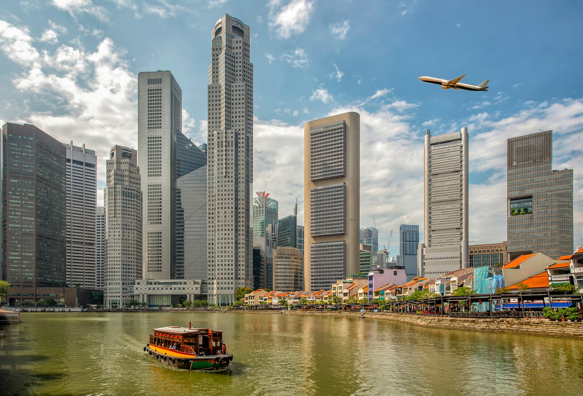 A plane flies over downtown Singapore's skyline with a ferry on the river below.