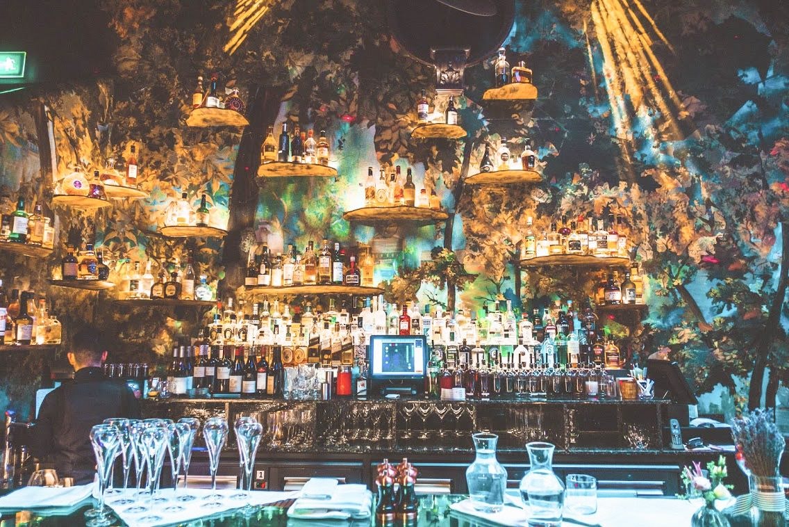 Interior of one of the bars at Sketch, Mayfair, London. The bar is decorated to look like an enchanted forest, with green walls, lots of plants and bottles nestled in amongst them. 
