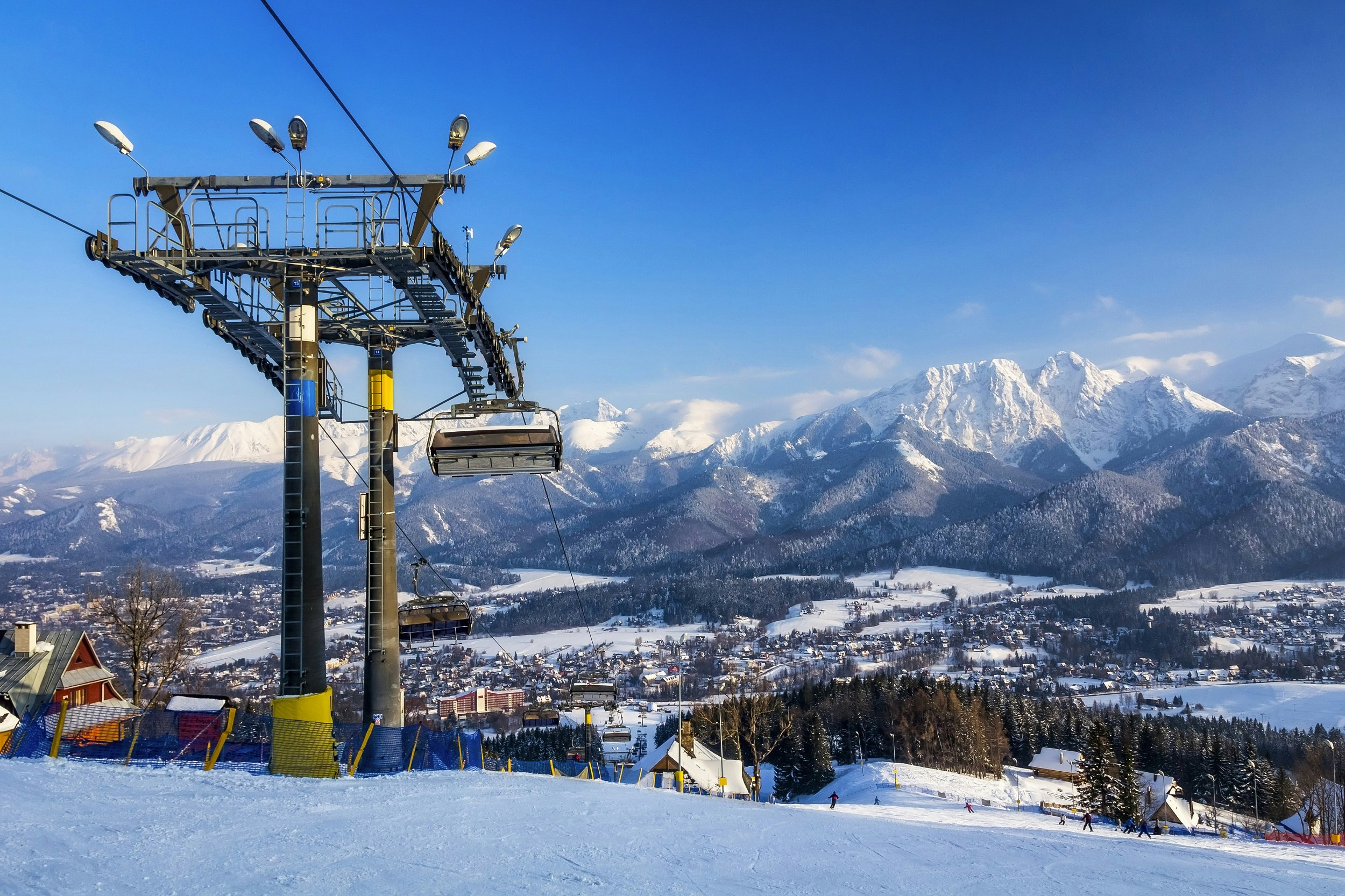 A ski lift on a snowy mountain with a range of snow-topped mountains stretching out in the distance. A town and woodland can be seen in the valley