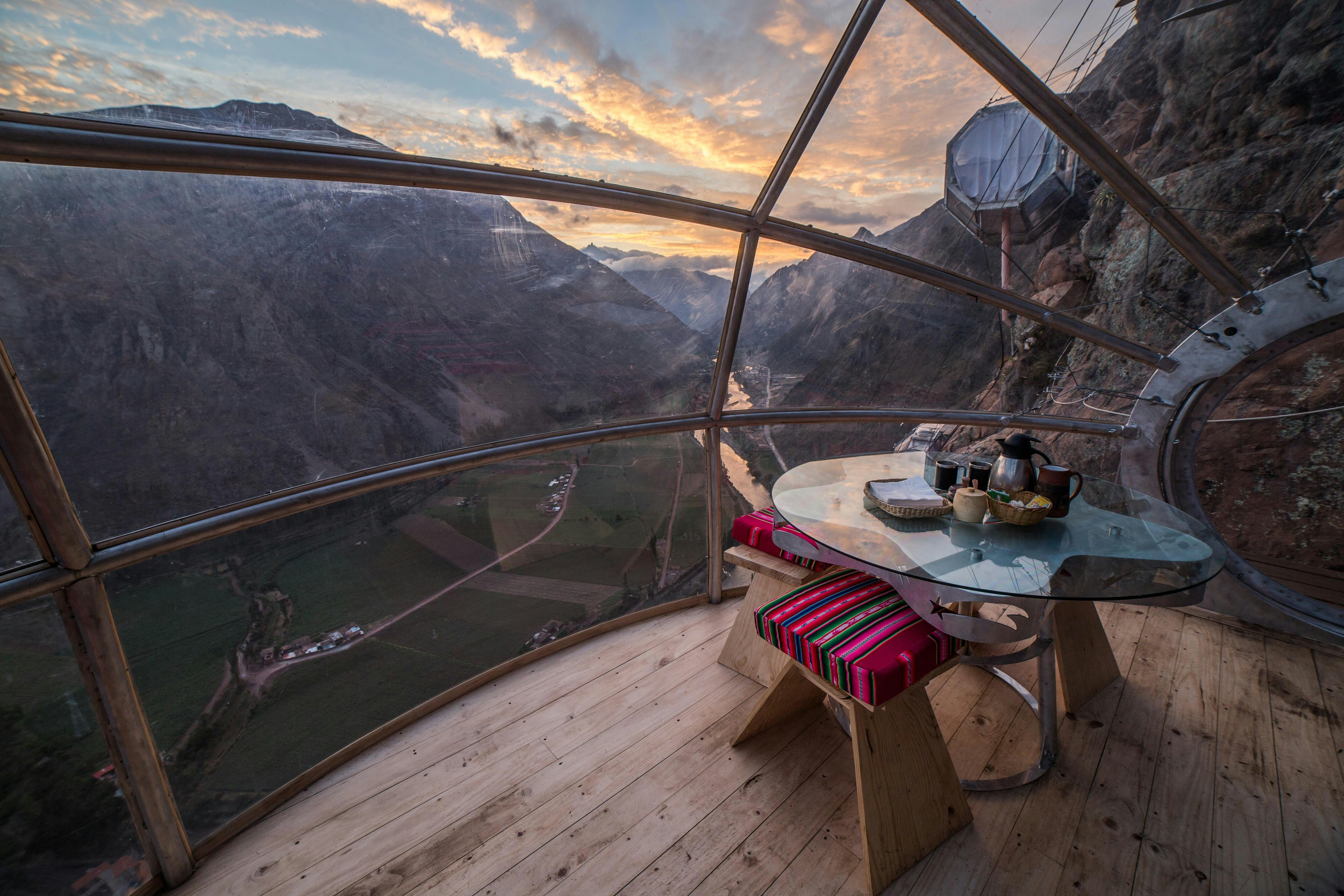 A table is set for coffee inside a glass structure with wooden floorboards on the floor. Through the glass we can see the structure is affixed to a cliff-face, and there's a view way down to the valley below and the huge mountains surrounding it.
