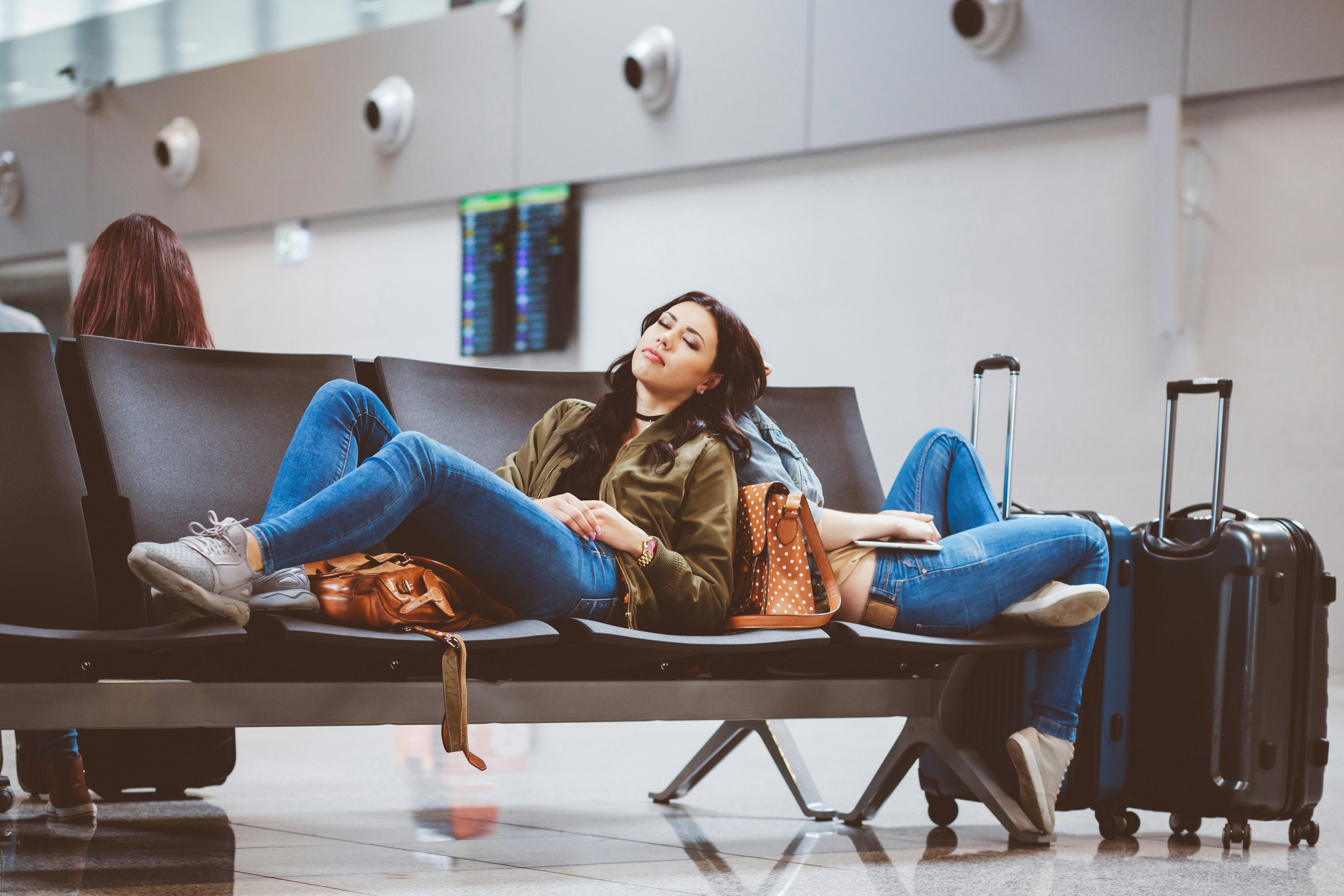 Two female travellers nap on a bench in an airport, propped up against their bags.