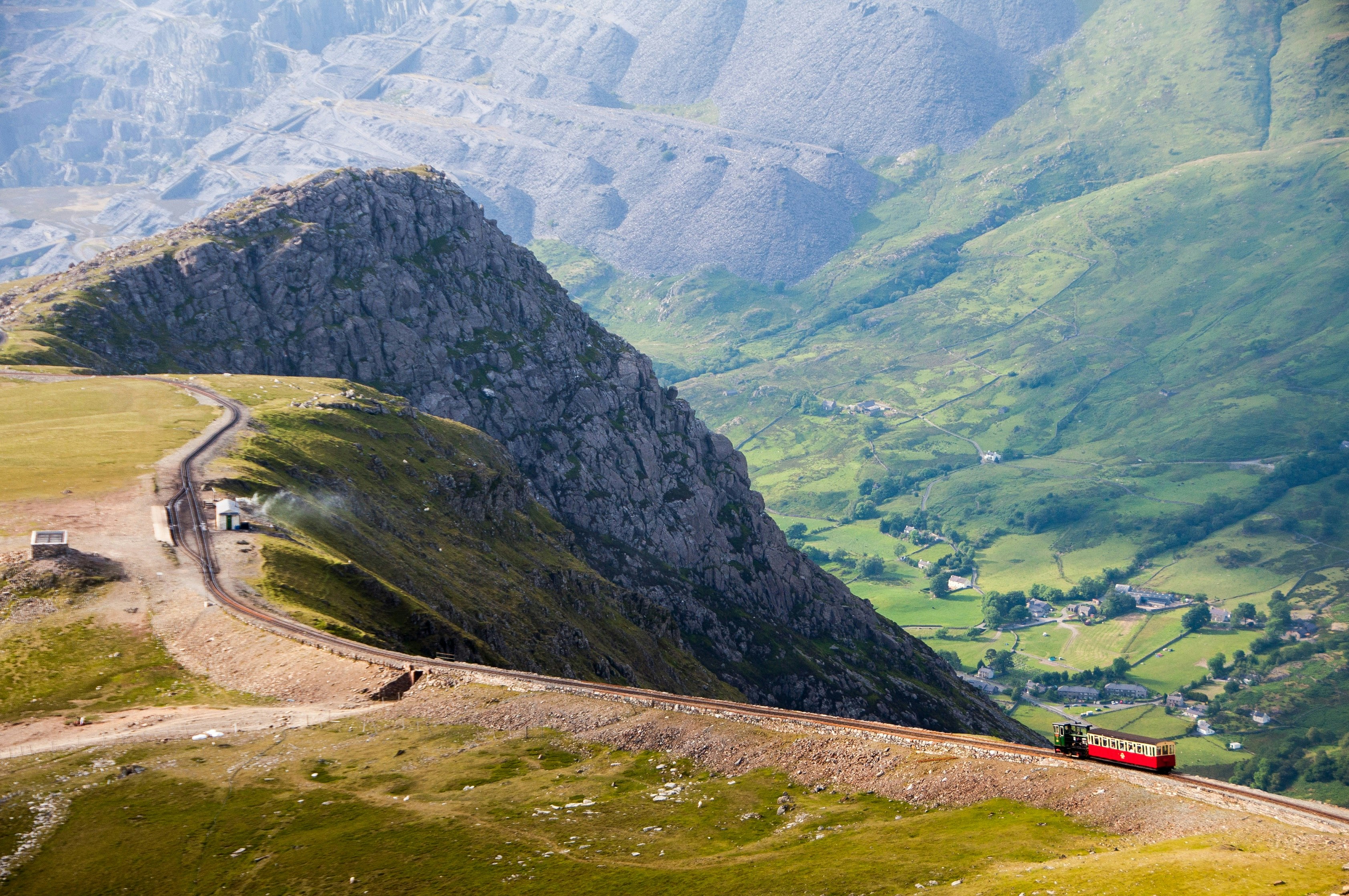 The Snowdon Mountain Railway running along the tracks on its way to the summit of Snowdon. The carriage is red, which stands out against the green hillsides. In the background, a small village is visible at the mountain's base.