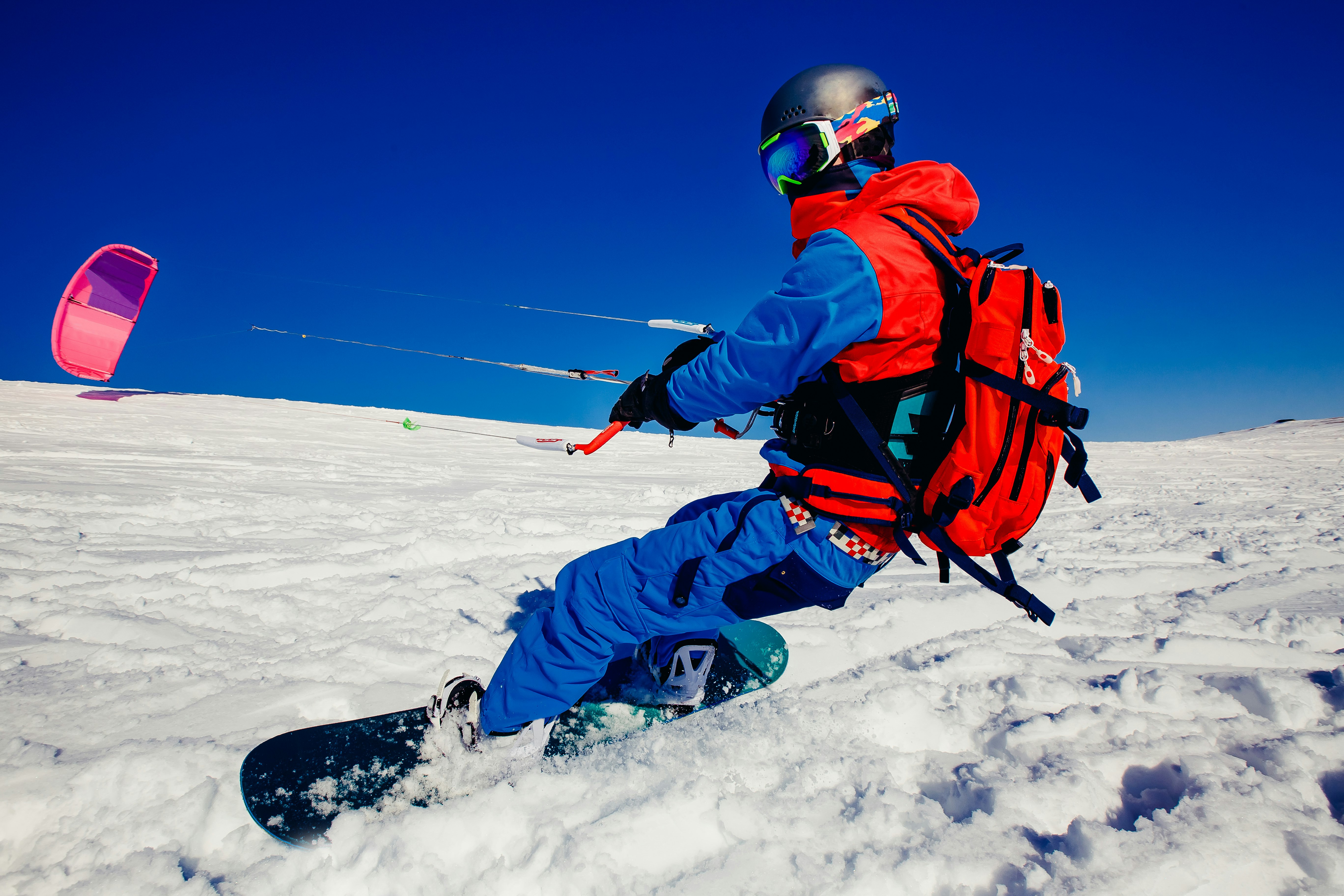 A person on a snowboard and wearing a bright blue and red snow outfit, googles and a helmet holds a handle attached to a large kit over snow