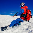 A person on a snowboard and wearing a bright blue and red snow outfit, googles and a helmet holds a handle attached to a large kit over snow