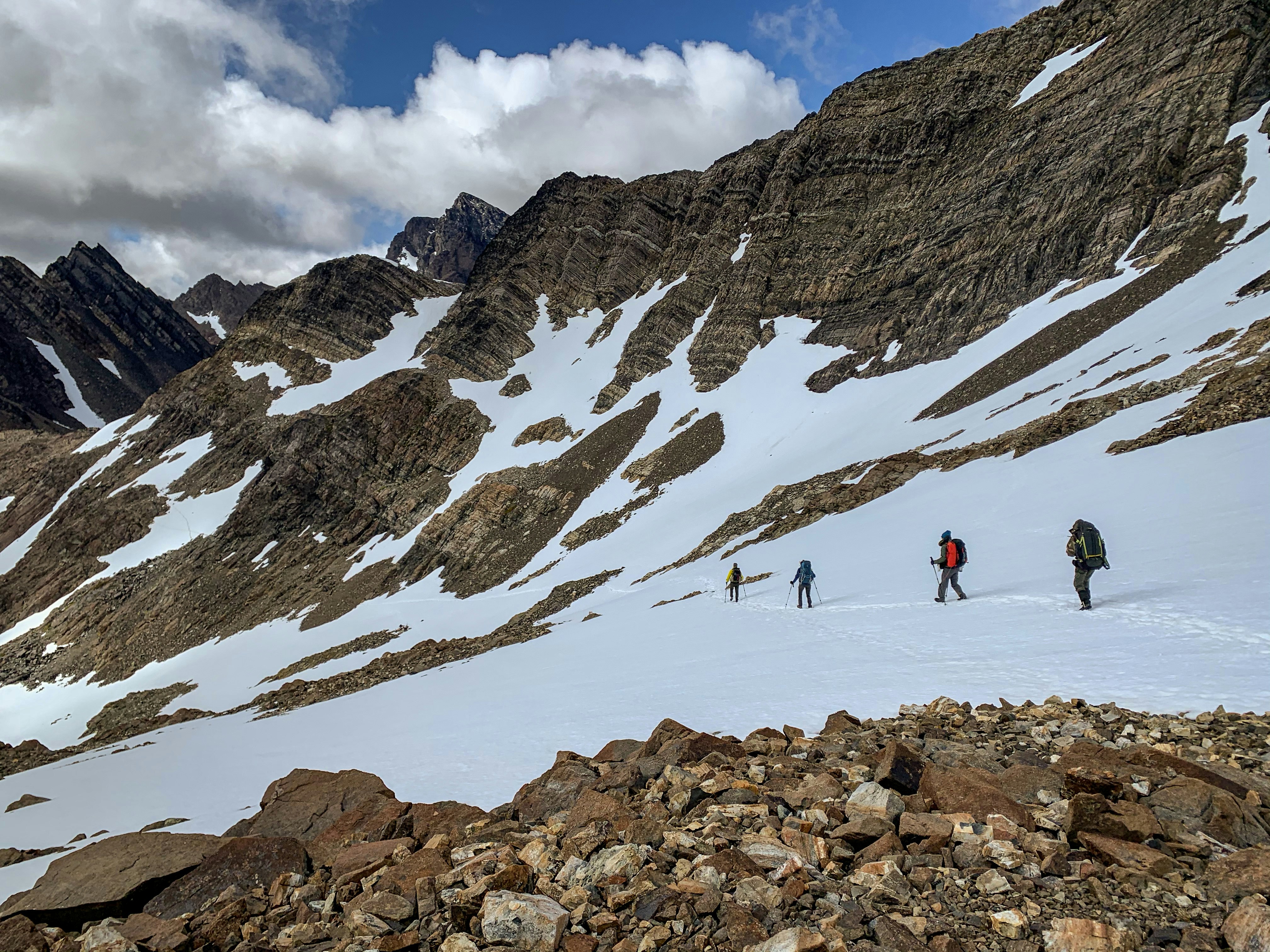 Four hikers with large backpacks trek across a snowy pass, with rocky peaks rising up the their right.