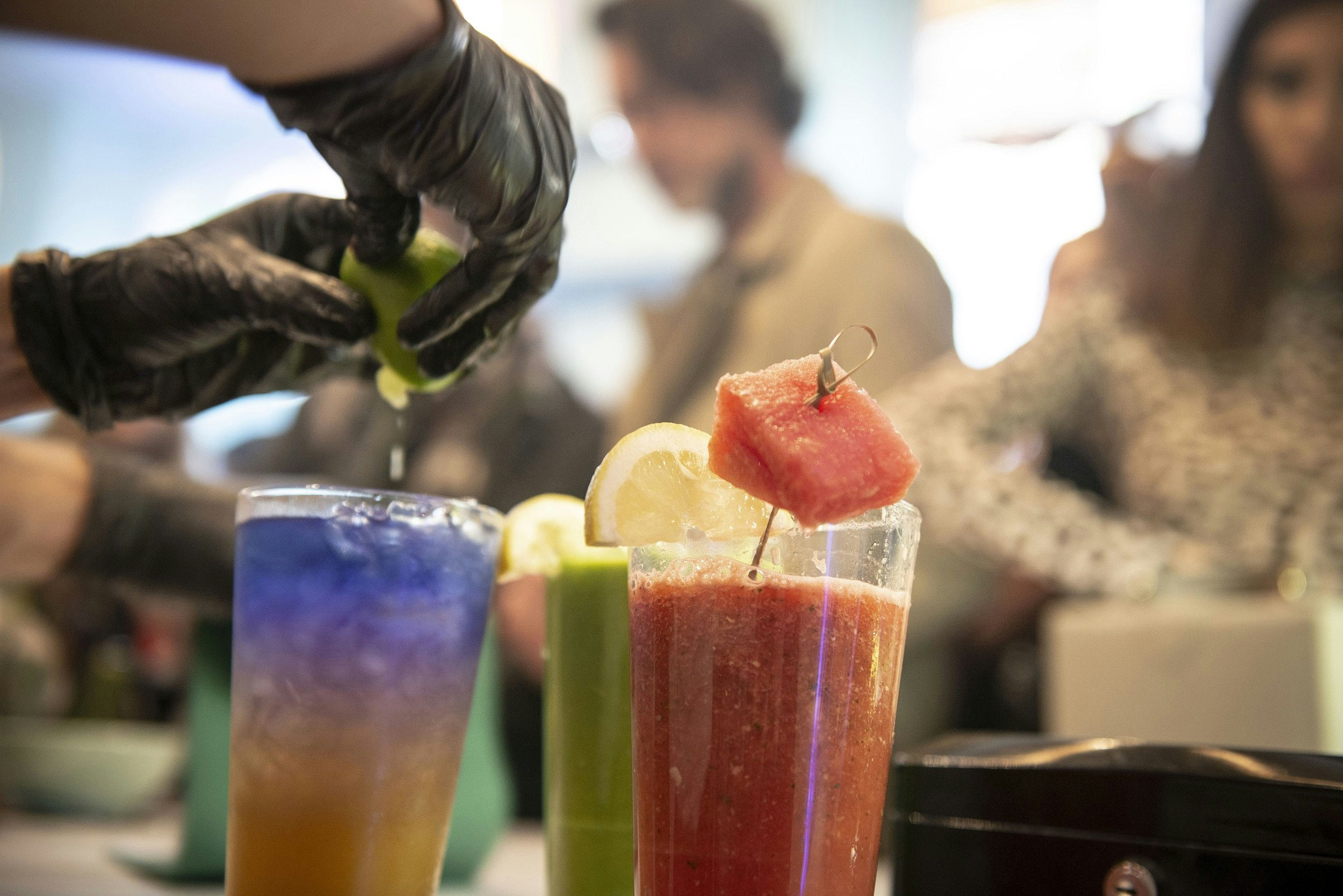 A close-up of a bartender's hands squeezing lime into a glass full of ice and a blue-and-orange drink. Next to it is a green drink and a red one topped with lemon and watermelon.