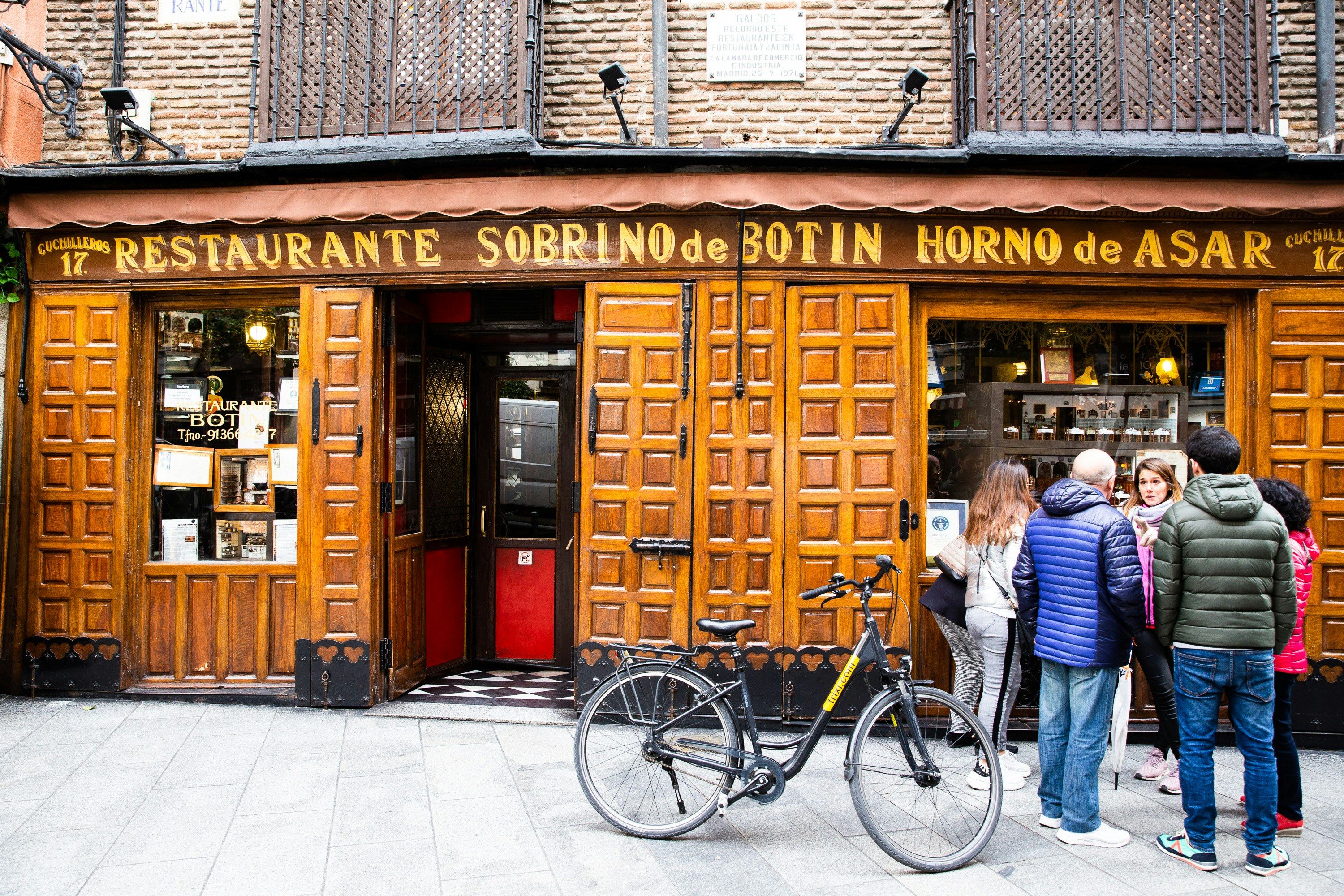 En grupp människor med en cykel utanför Sobrino de Botín, som har en träpanel och en brun skylt med restaurangens namn med gul skrift. 
