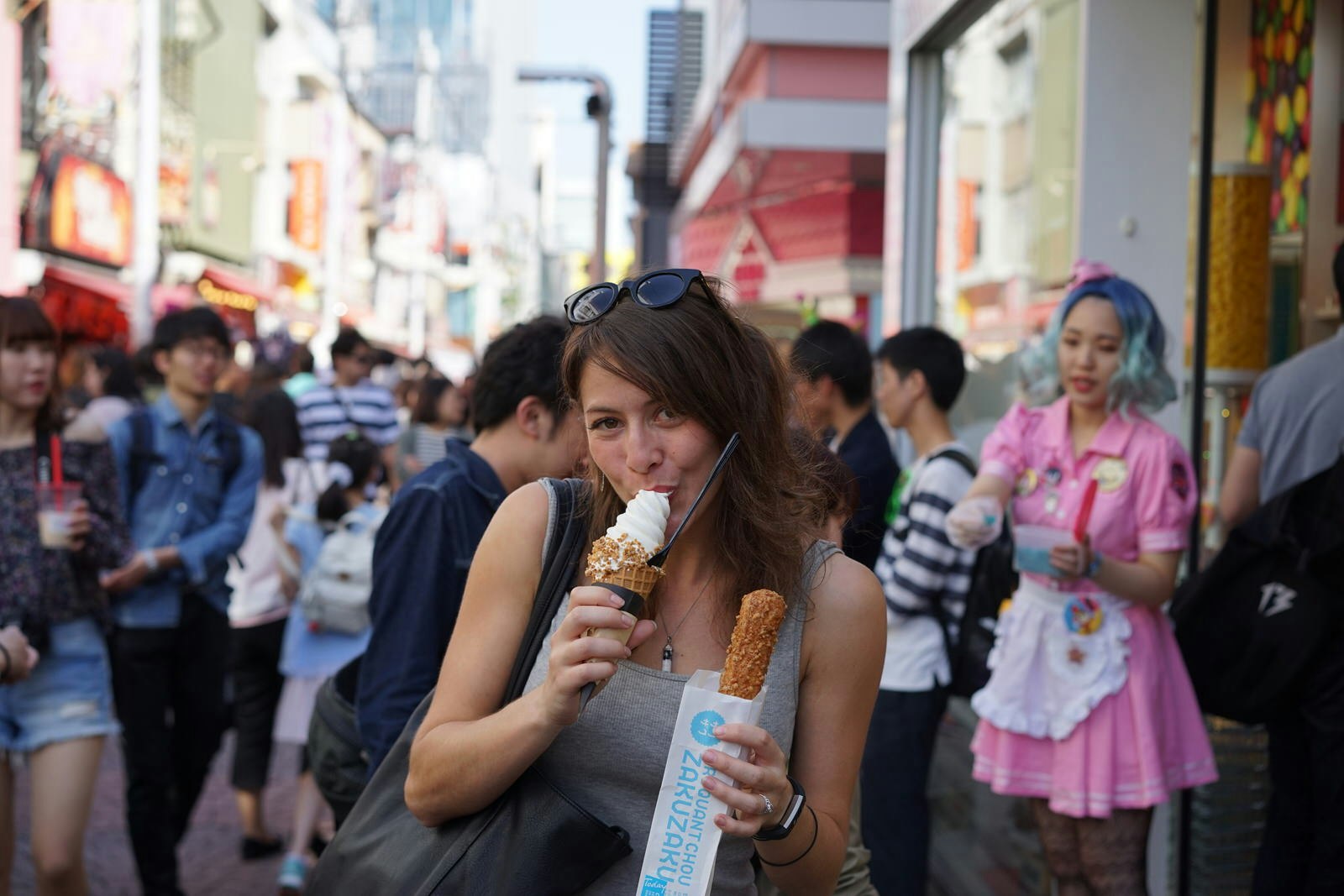 A woman eating an ice cream cone while holding a churro in her other hand in the middle of a busy walkway in Tokyo. 