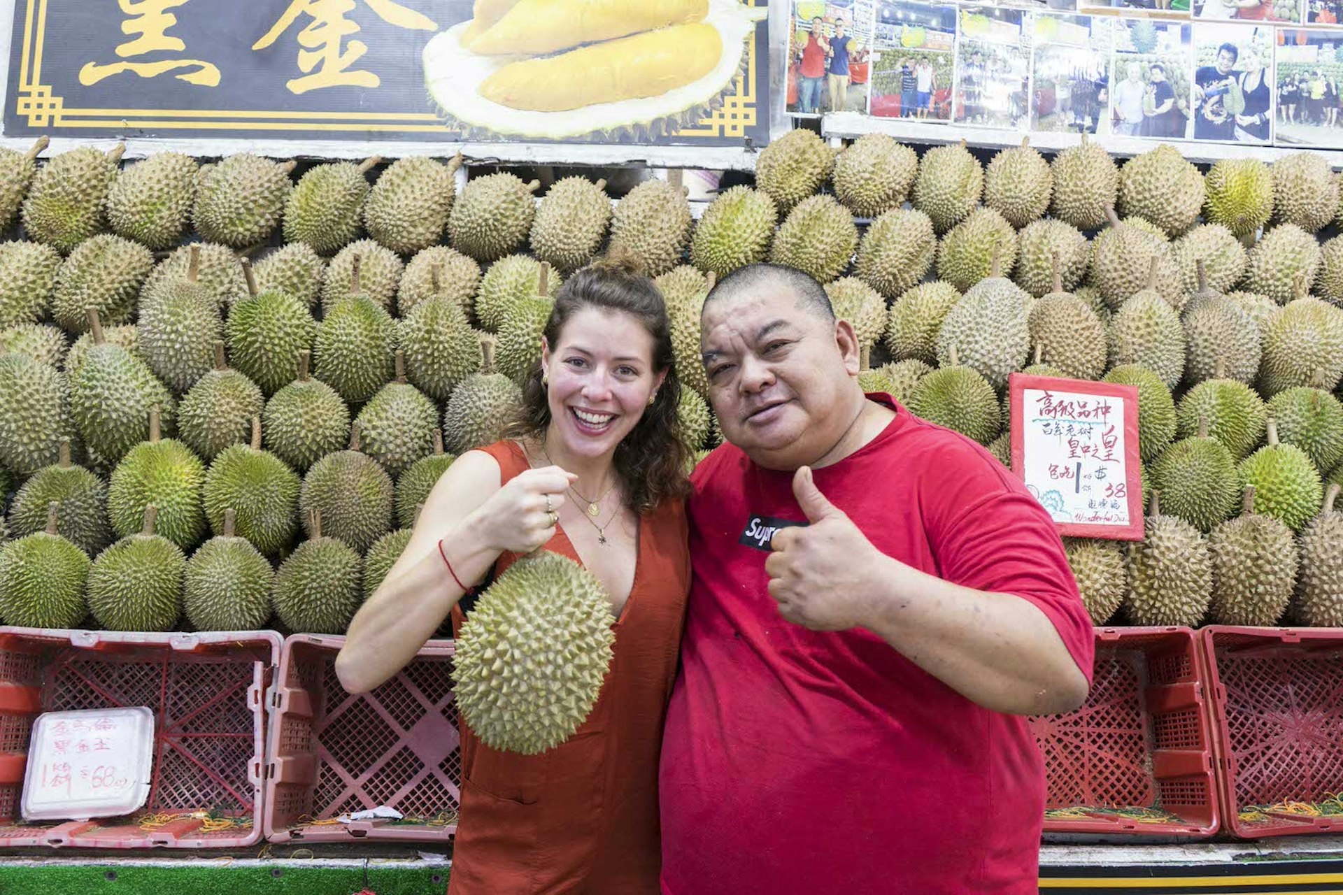 A woman smiling and holding a large durian while embracing a man in a red t-shirt with his thumb up. They are standing in front of a stall jam-packed with durians. 