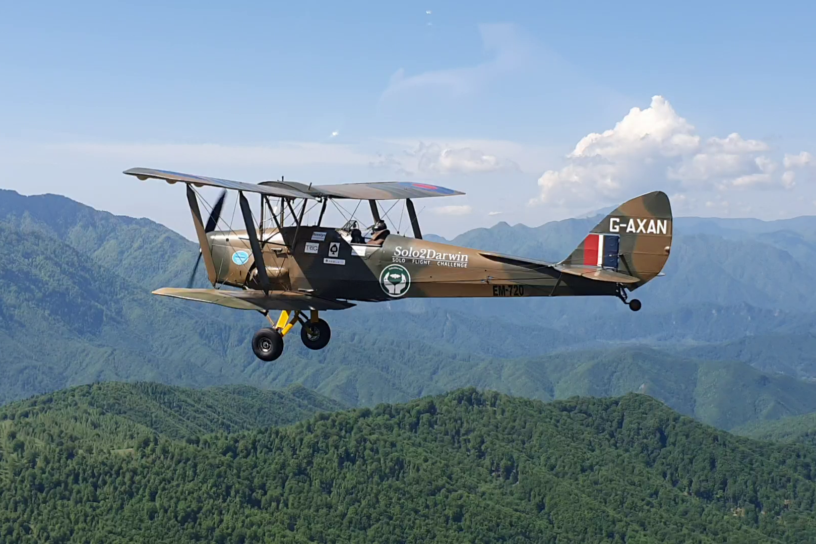 A Tiger Moth plane with 'Solo2Darwin' on the side flies over lush green hillsides
