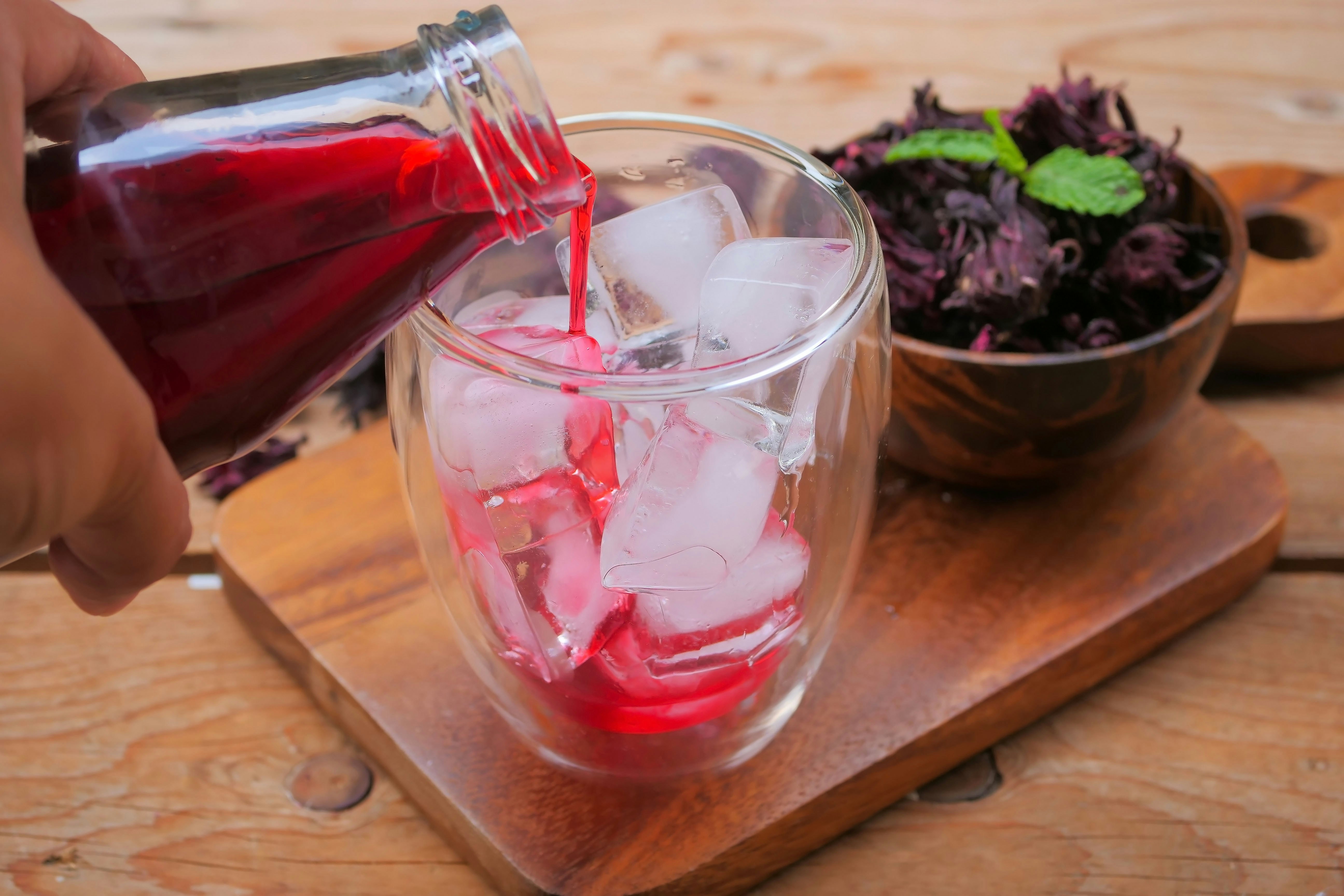 A person pours crimson-colored sorrel into a glass filled with ice. In the background is a bowl of dried sorrel topped with a green herb.