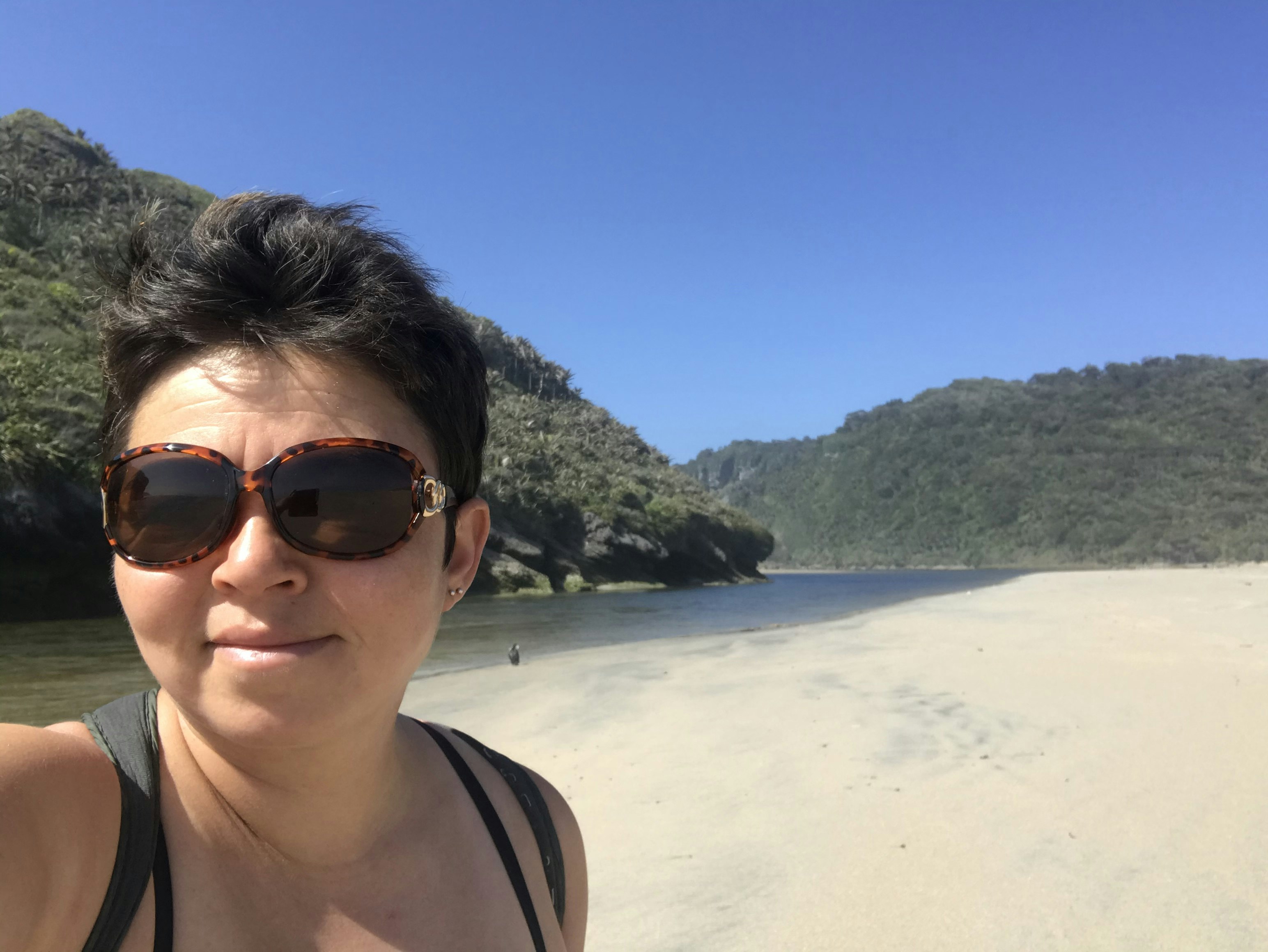 Anna Kaminski takes a selfie with a river, beach and green hills behind her.